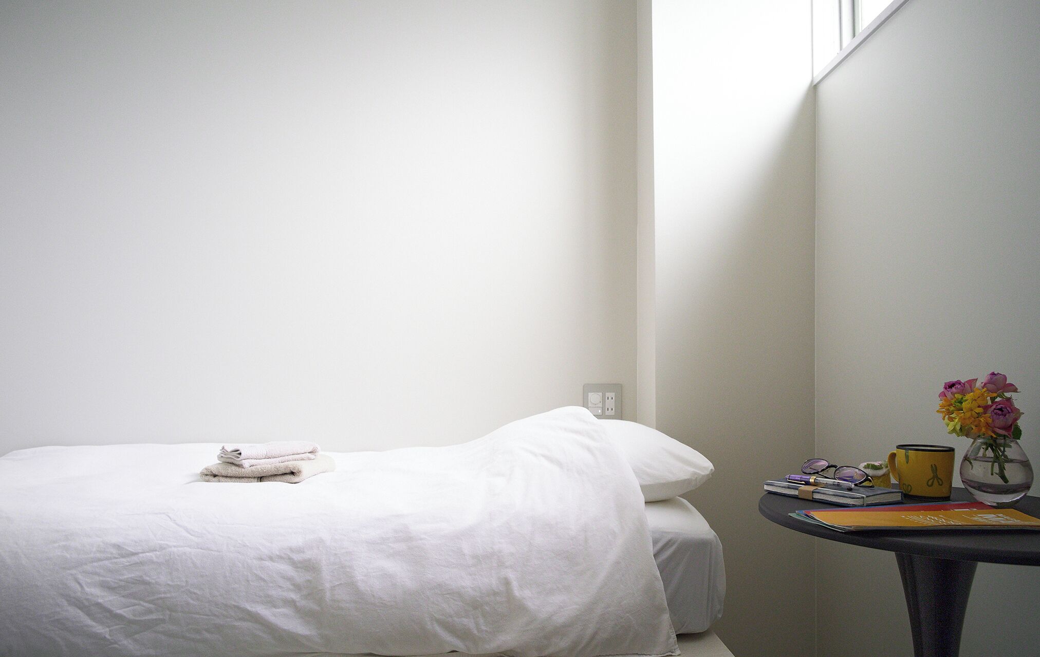 A neatly made bed with a folded towel, a small table with a vase of flowers, and a window in the background.