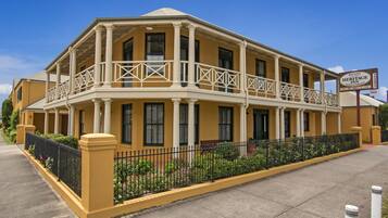 Un edificio amarillo de dos pisos con un balcón, un cartel que dice Ballina Heritage Inn y una valla negra adornada con flores.