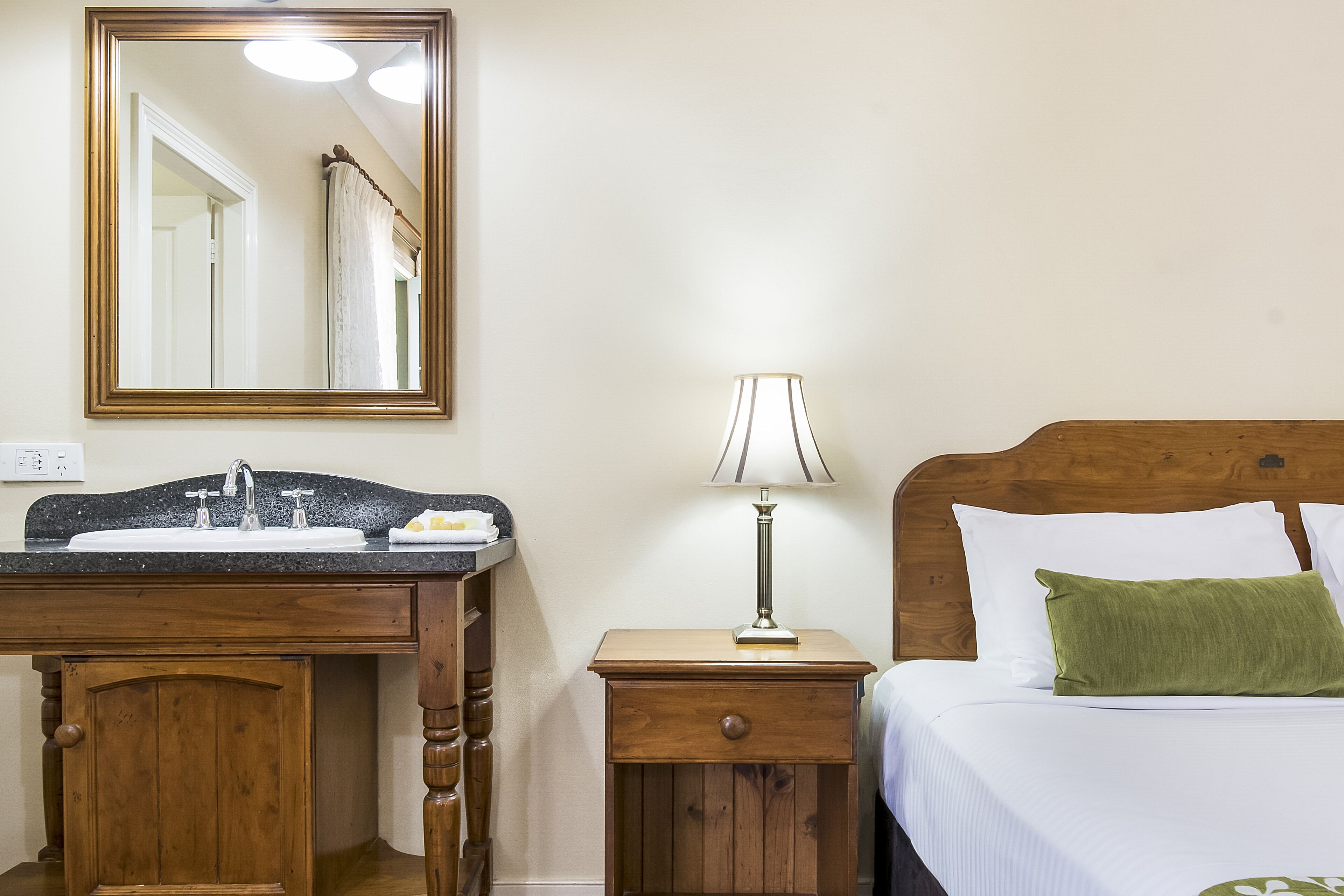 A hotel room with a wooden cabinet with a sink, a bedside table with a lamp, and a bed with pillows.