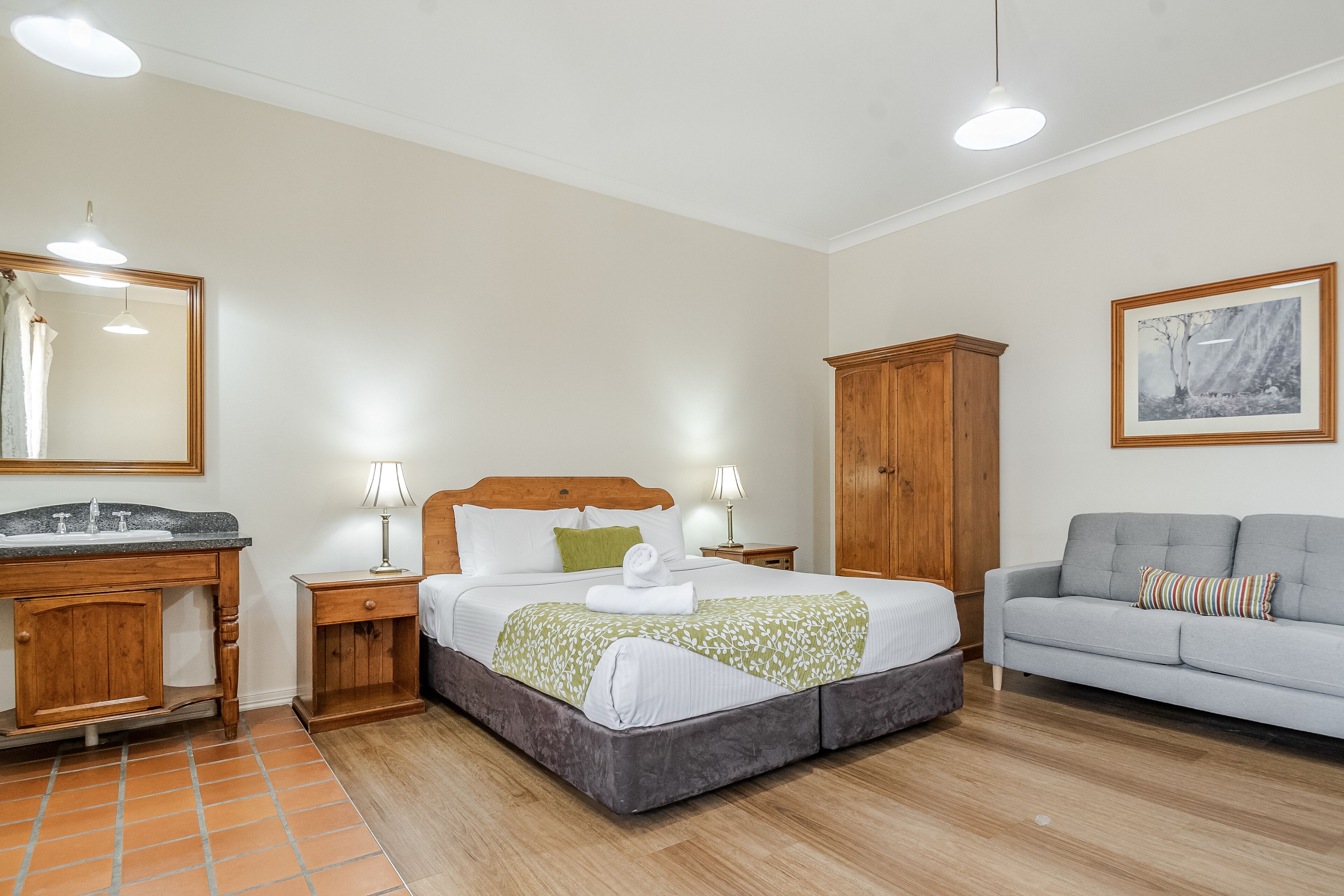 Un dormitorio con cama, mesitas de noche, un armario de madera, un sofá gris y un cuadro en la pared.