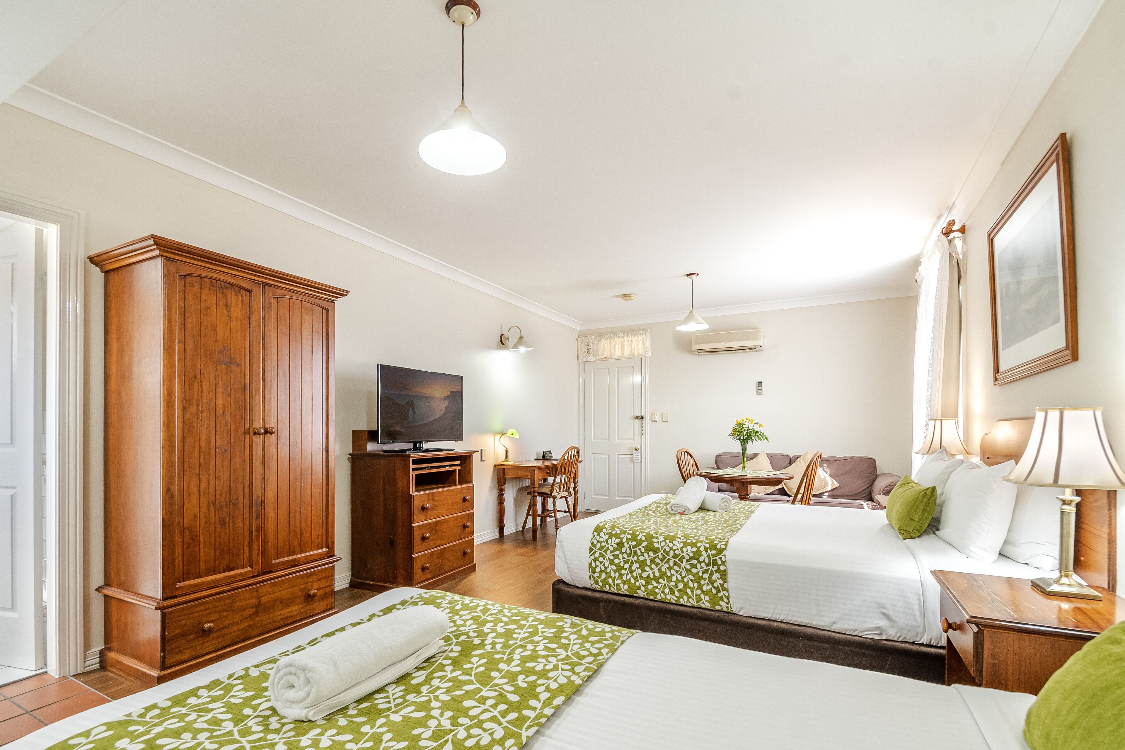 A hotel room with two beds, a wooden wardrobe, a desk with a chair, a television, and a dining area with a table and chairs.
