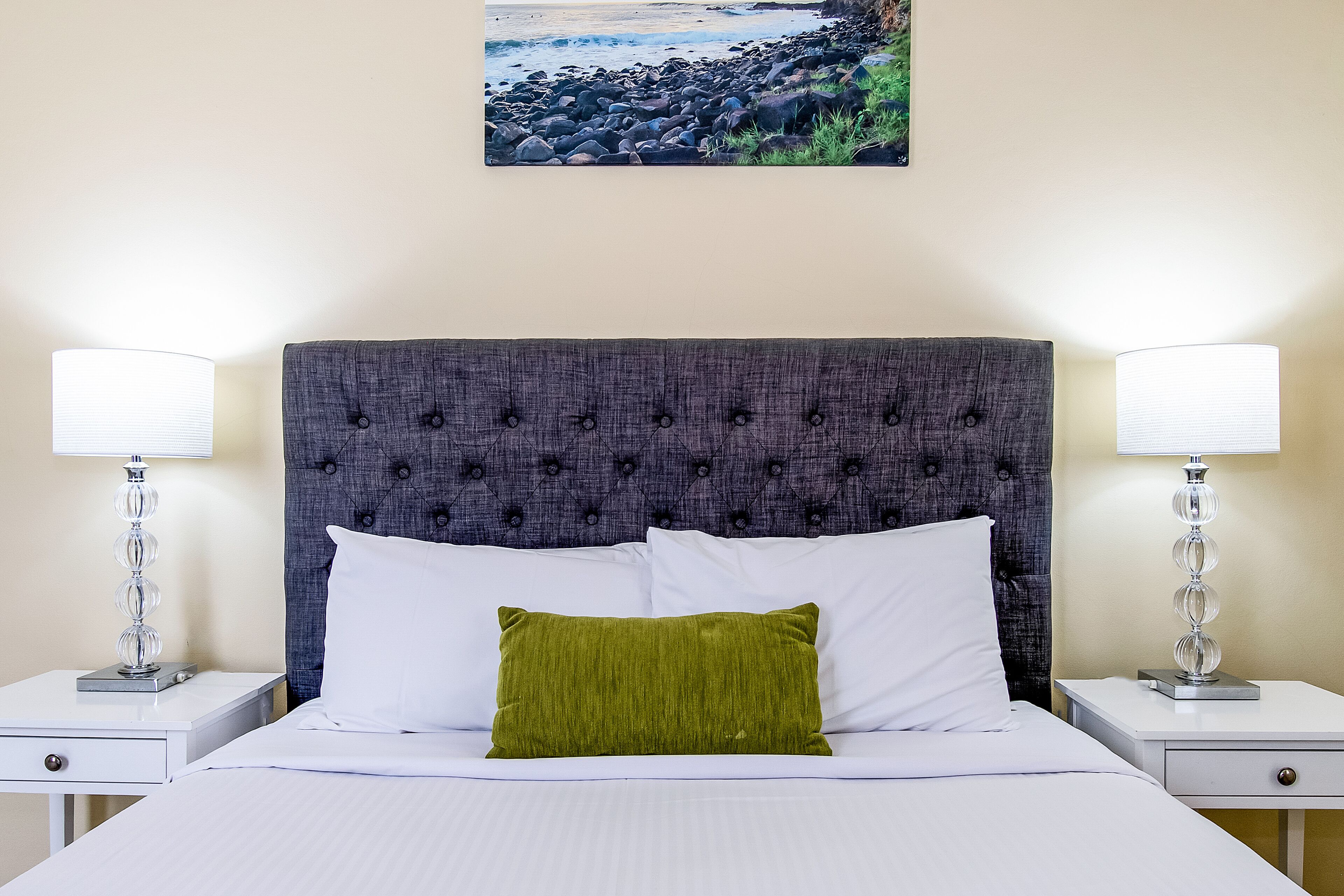A neatly made bed with white linens and a green pillow, flanked by two bedside tables with lamps, against a wall with a framed landscape painting.
