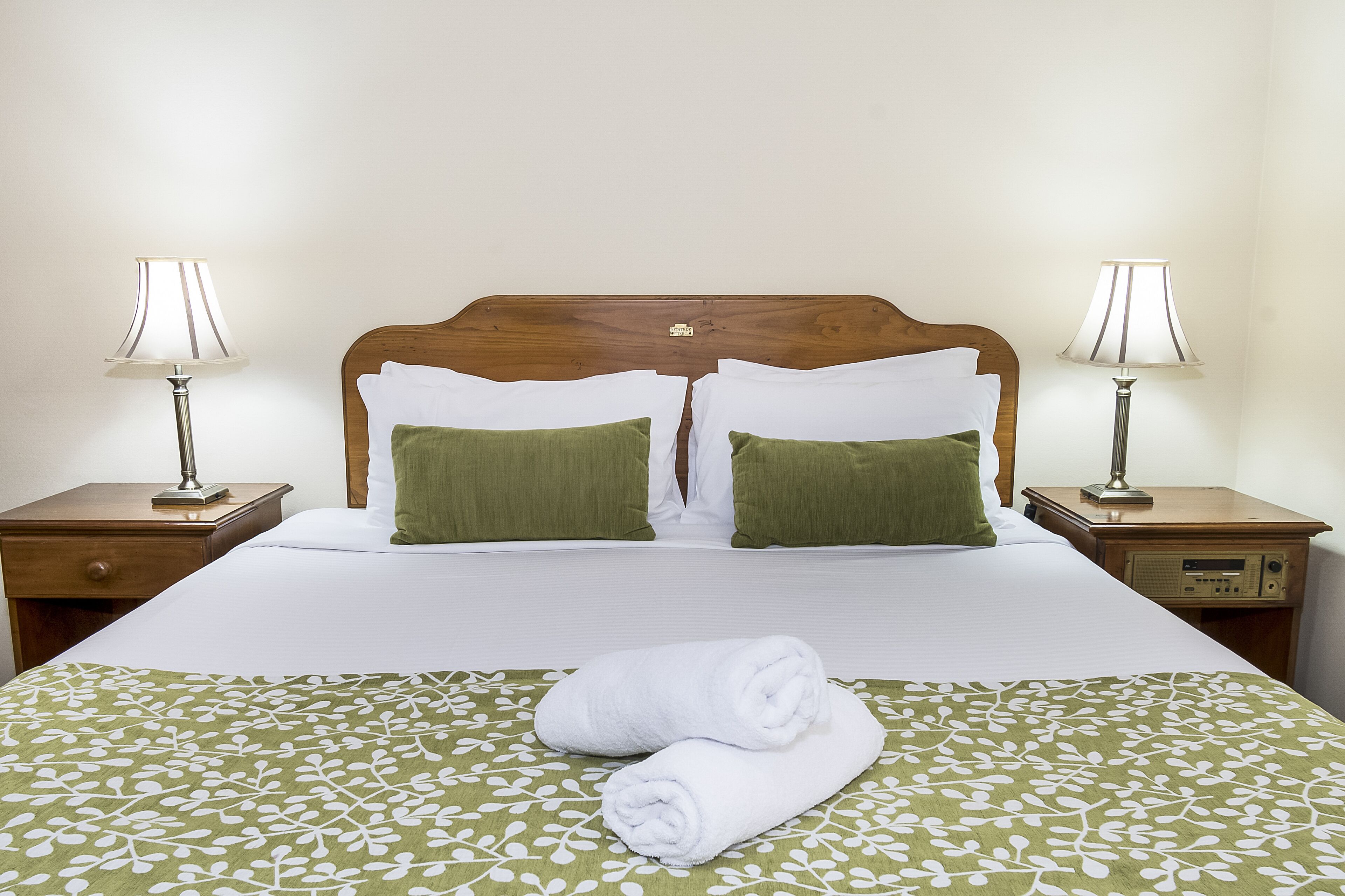 Una cama bien hecha con una colcha verde estampada, dos almohadas blancas y un par de toallas blancas dobladas.