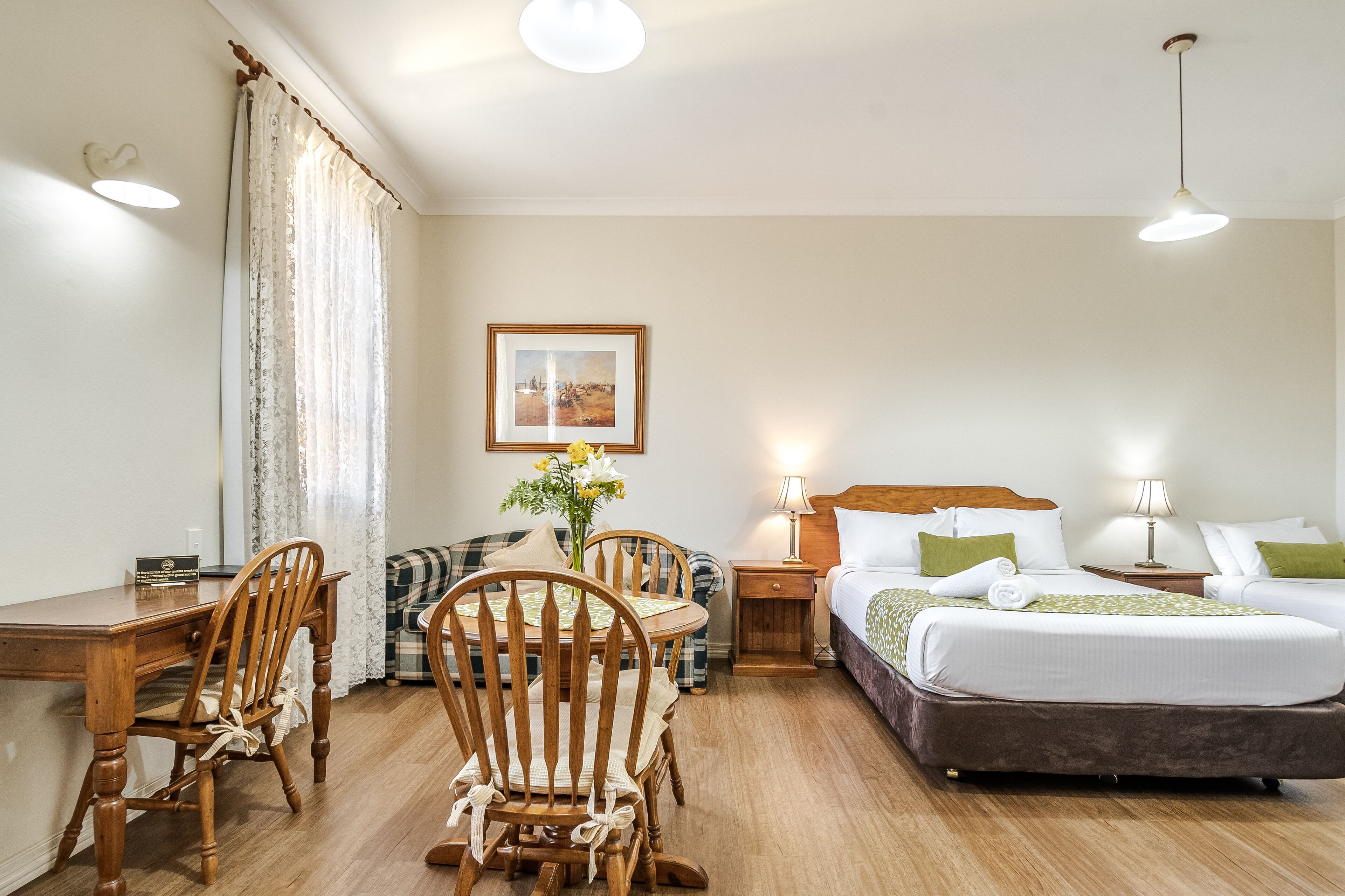 Une chambre d’hôtel avec un bureau en bois, deux chaises, un lit avec des oreillers verts, une table de chevet avec une lampe et un tableau encadré au mur.