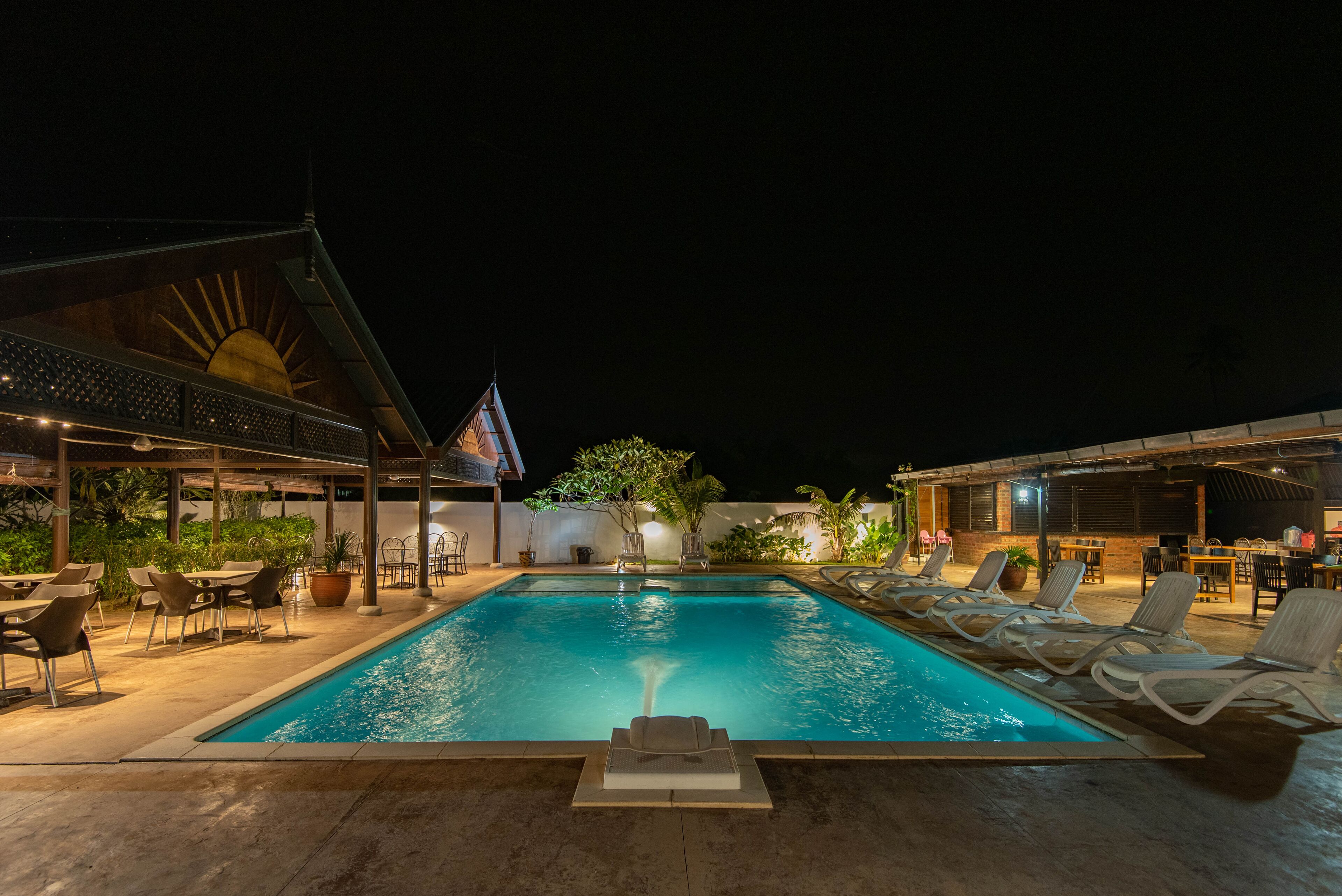 A swimming pool with lounge chairs and tables, surrounded by potted plants and a covered area with a sun motif.