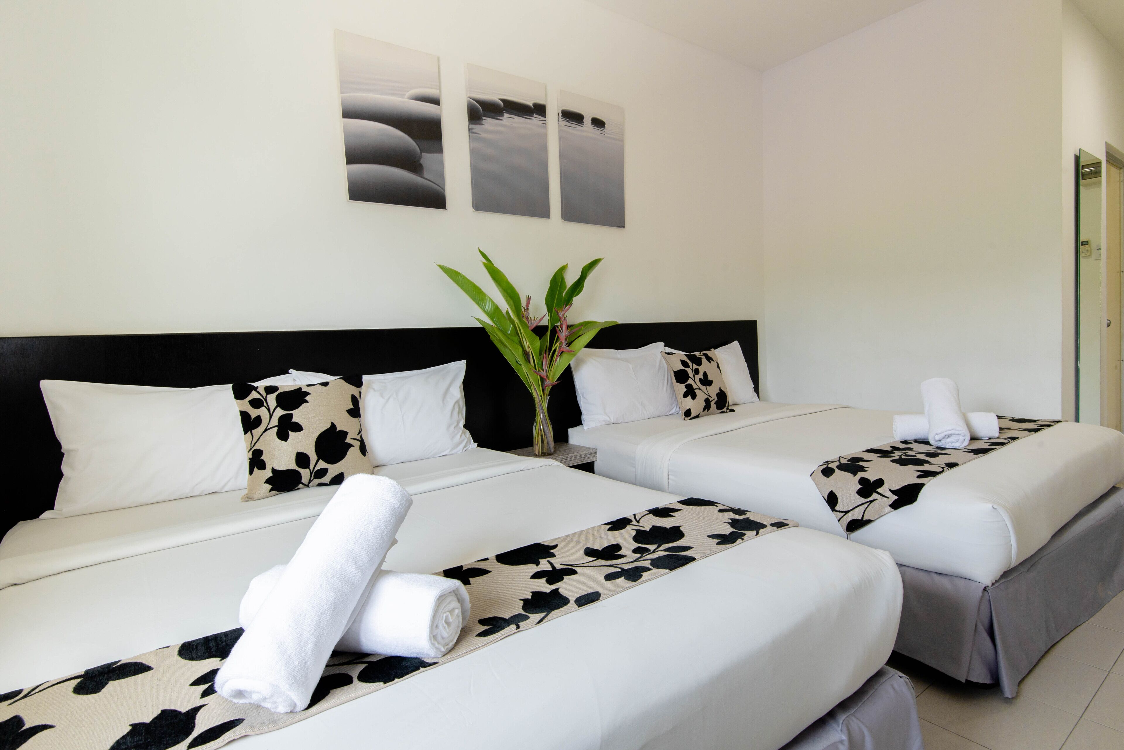 Habitación de hotel con dos camas, ropa de cama blanca, almohadas florales en blanco y negro, una planta y tres cuadros en la pared.