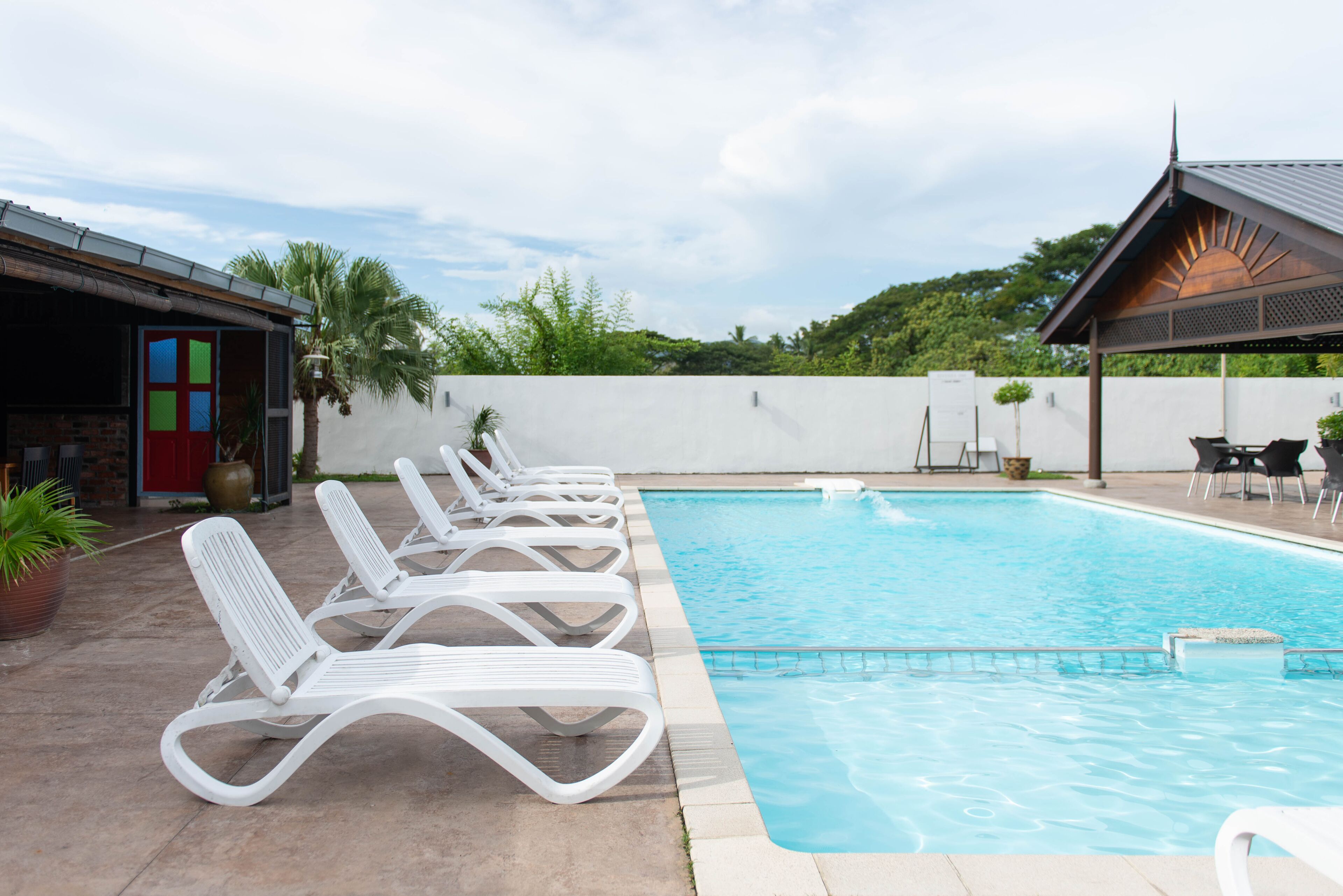 A swimming pool with white lounge chairs, a covered seating area, and a view of greenery in the background.