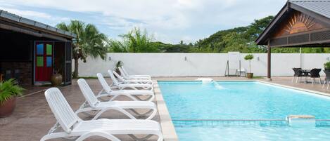 A swimming pool with white lounge chairs, a covered seating area, and a view of greenery in the background.