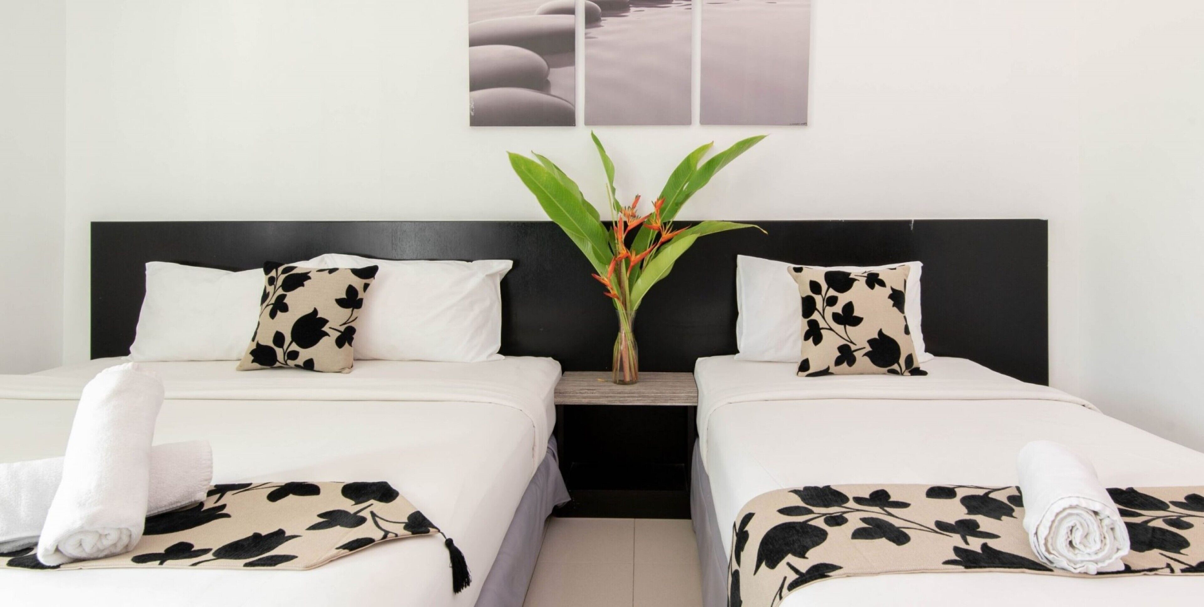 A hotel room with two beds, each with a black and white patterned pillow, a white bedspread, and a rolled white towel on the bed. There is a nightstand with a vase of flowers between the beds.