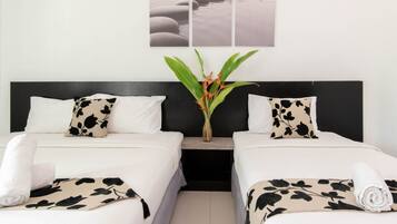 A hotel room with two beds, each with a black and white patterned pillow, a white bedspread, and a rolled white towel on the bed. There is a nightstand with a vase of flowers between the beds.