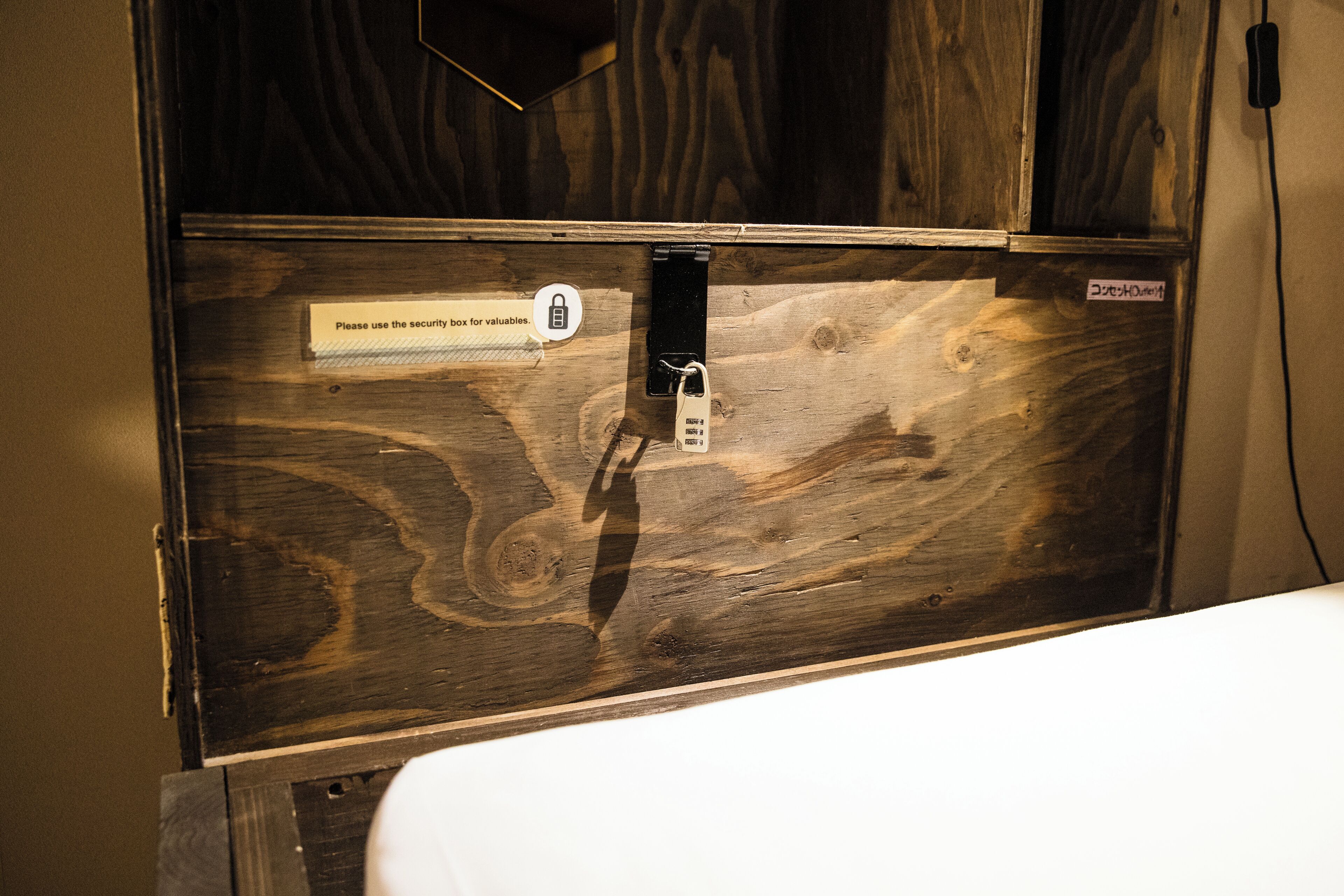 Una caja de seguridad de madera con un candado y llave, un letrero que indica usarla para objetos de valor, y un enchufe en la pared.