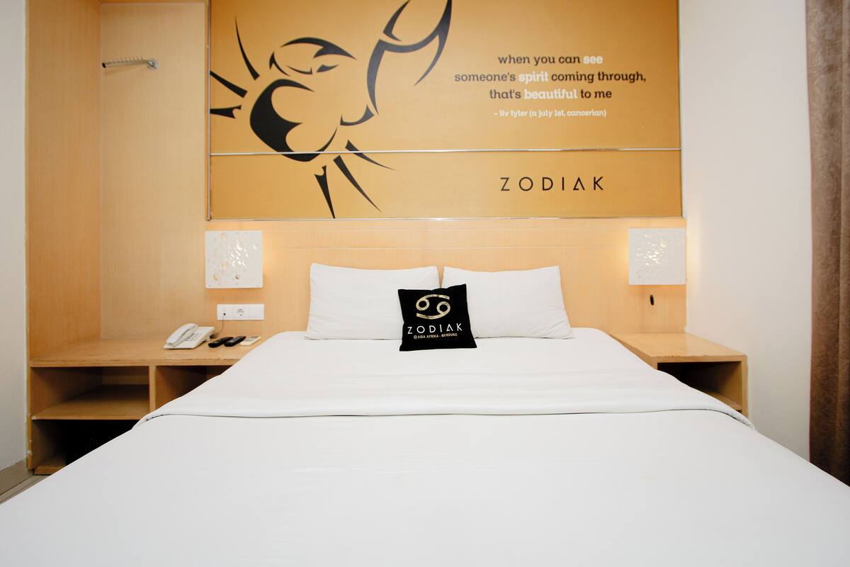 A hotel room with a large bed, two bedside tables, a wall with a zodiac sign and quote, and a small shelf with a phone and remote.