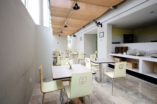 A modern dining area with multiple tables and chairs, a kitchen in the background, and a view of the outside.