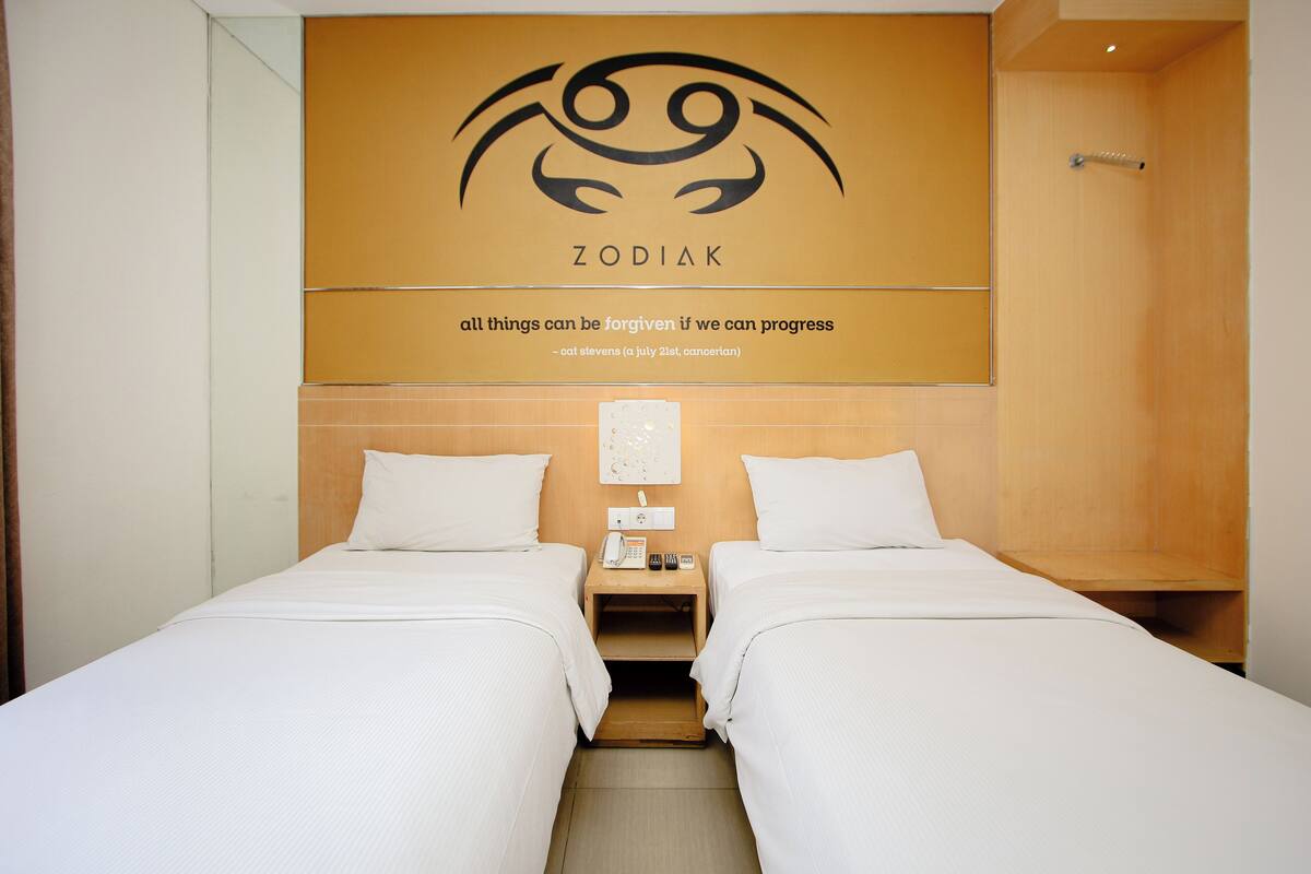 A hotel room with two beds, a wooden headboard, a wall with a zodiac sign and a quote, and a bedside table with a phone and remote controls.