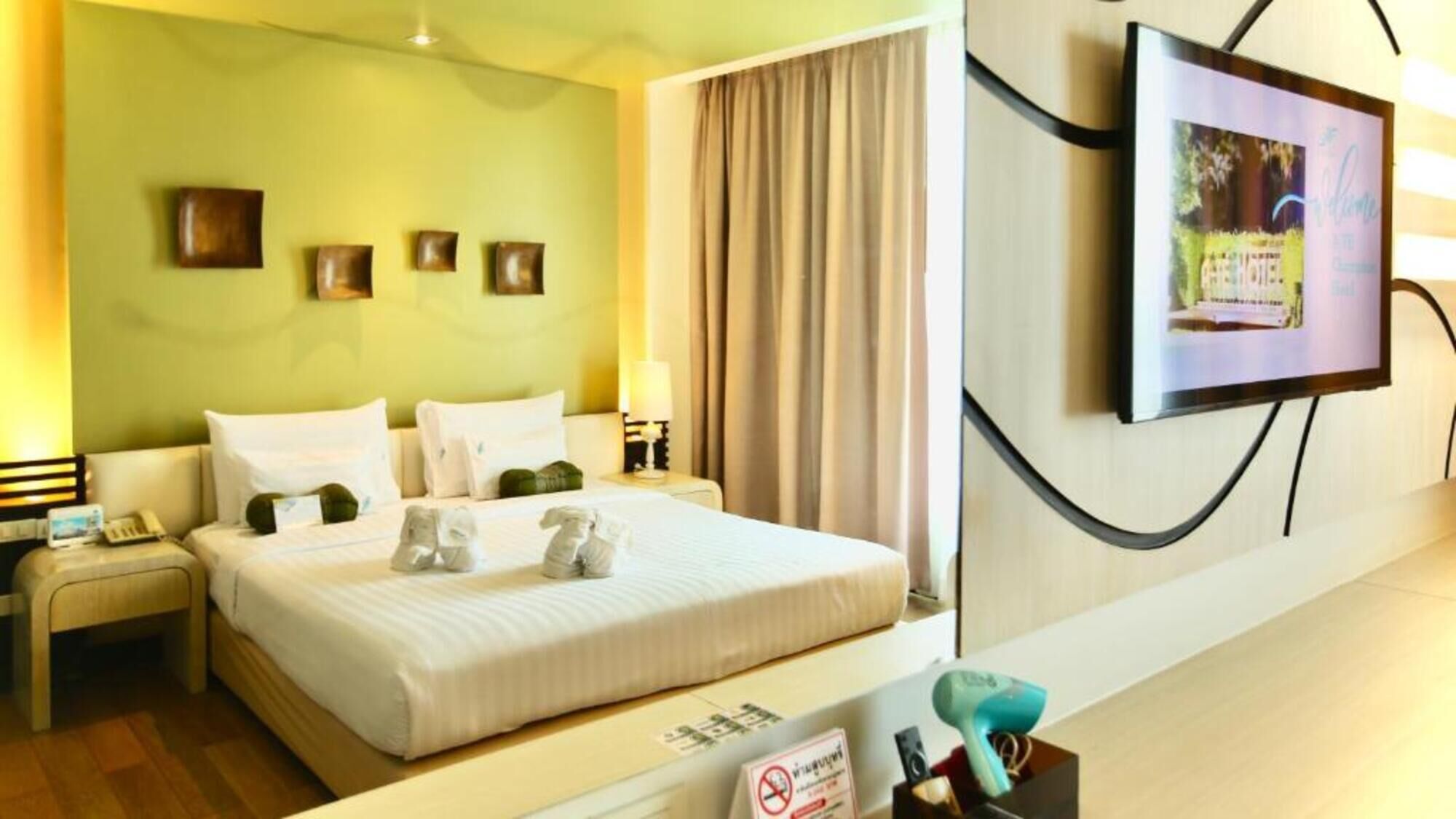 A hotel room with a bed, a nightstand, a lamp, a TV on the wall, and a sign with a no-smoking symbol.