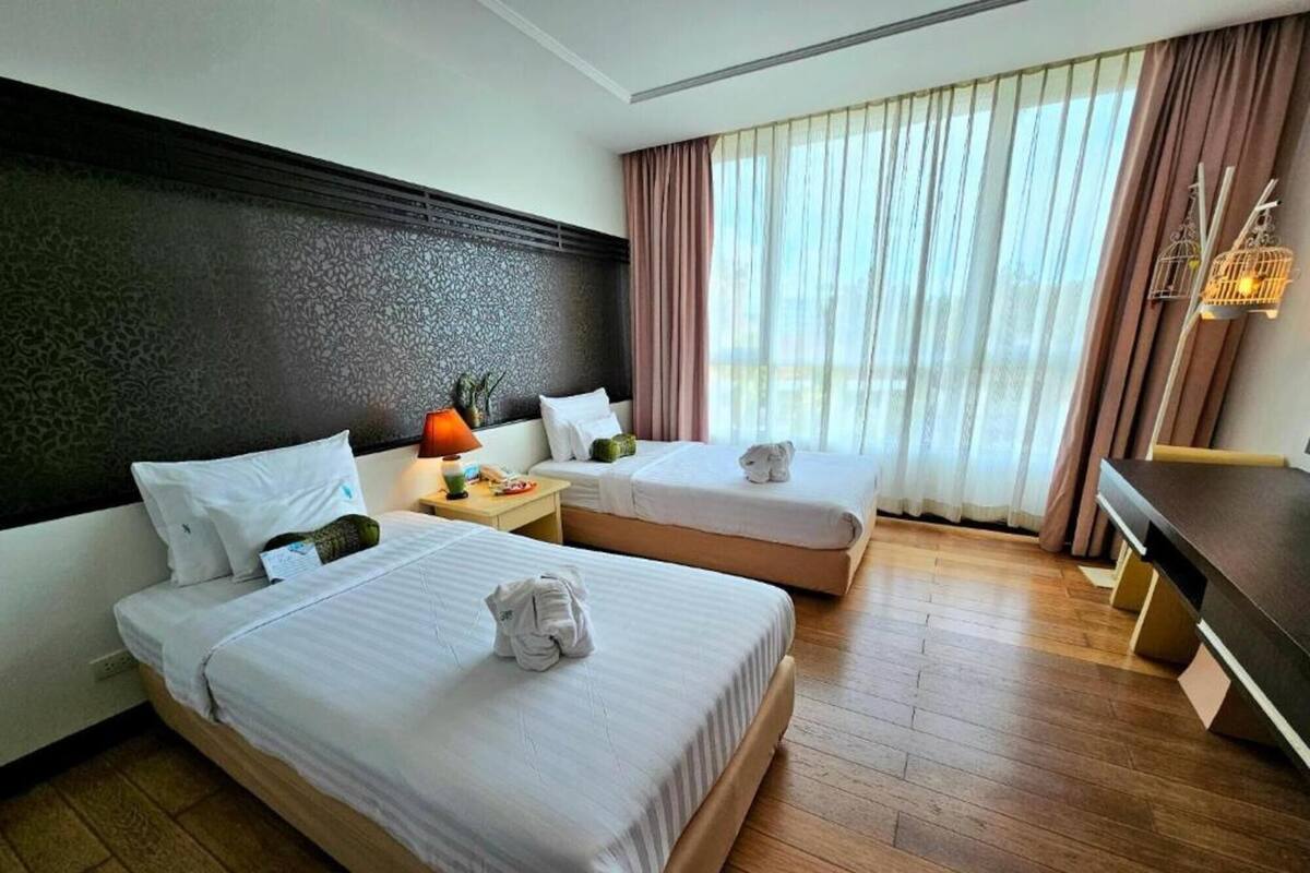 A hotel room with two beds, a nightstand with a lamp, a wooden dresser, and a window with curtains.