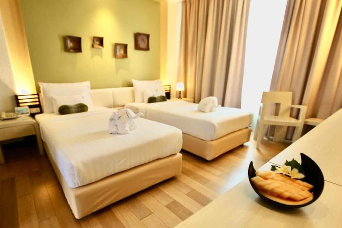 A hotel room with two beds, a small table with a fruit bowl, a chair, and a nightstand with a lamp.