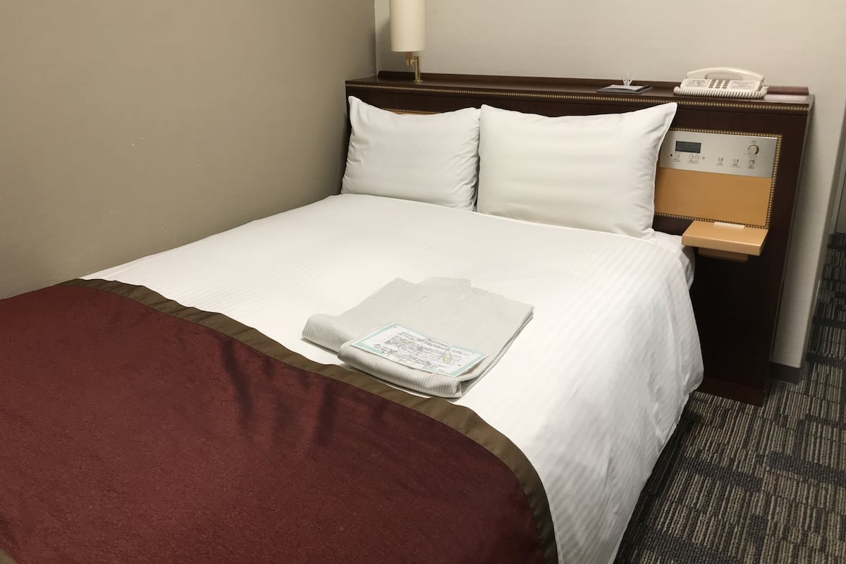 A hotel room with a neatly made bed, a bedside table with a lamp, a telephone, and a small shelf.