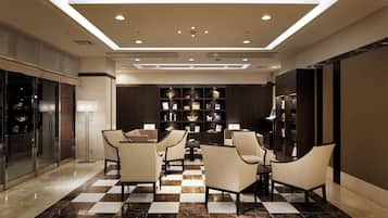 Um hall de hotel com piso xadrez, uma área de estar central com cadeiras, uma estante de livros e um design de teto moderno.