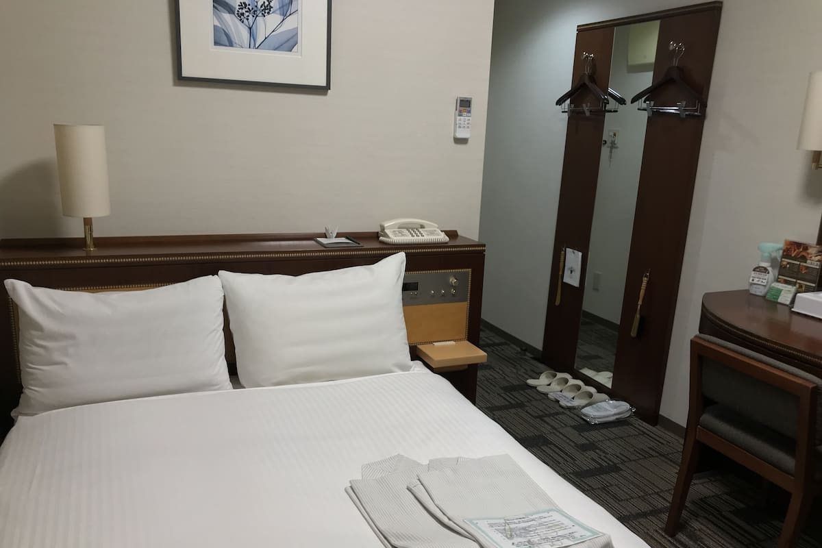 A hotel room with a neatly made bed, a bedside telephone, a mirror, a coat rack, and a small table with toiletries.