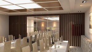 A modern dining area with a long table set for a meal, surrounded by chairs, and a sofa in the corner. - Bella Hotel Surabaya (Surabaya)