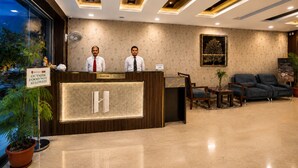 A hotel reception area with a reception desk, two receptionists, a sign prohibiting outside food, a reception sign, a large H-shaped logo, and a seating area with a sofa and chairs. - Horizon Hotel (Udaipur)