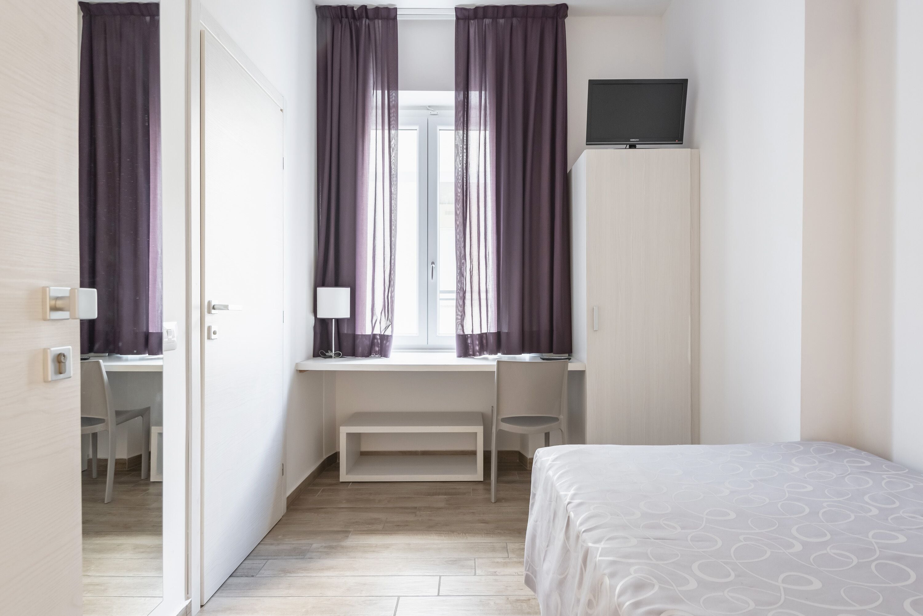 Habitación de hotel con cama, escritorio con silla, televisor en la pared y ventana con cortinas.