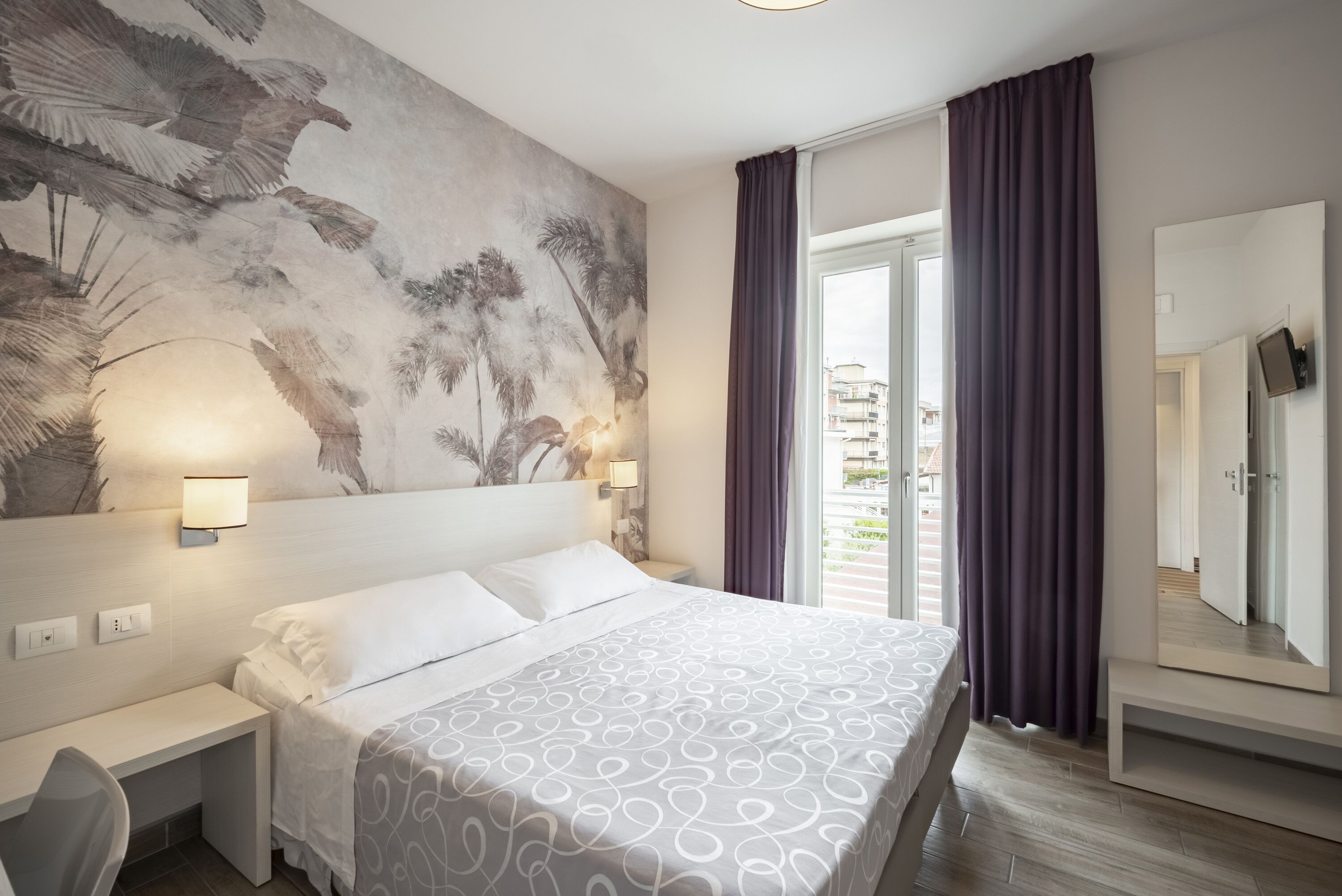 A modern hotel room with a large bed, a desk, a wall-mounted TV, and a balcony with curtains.