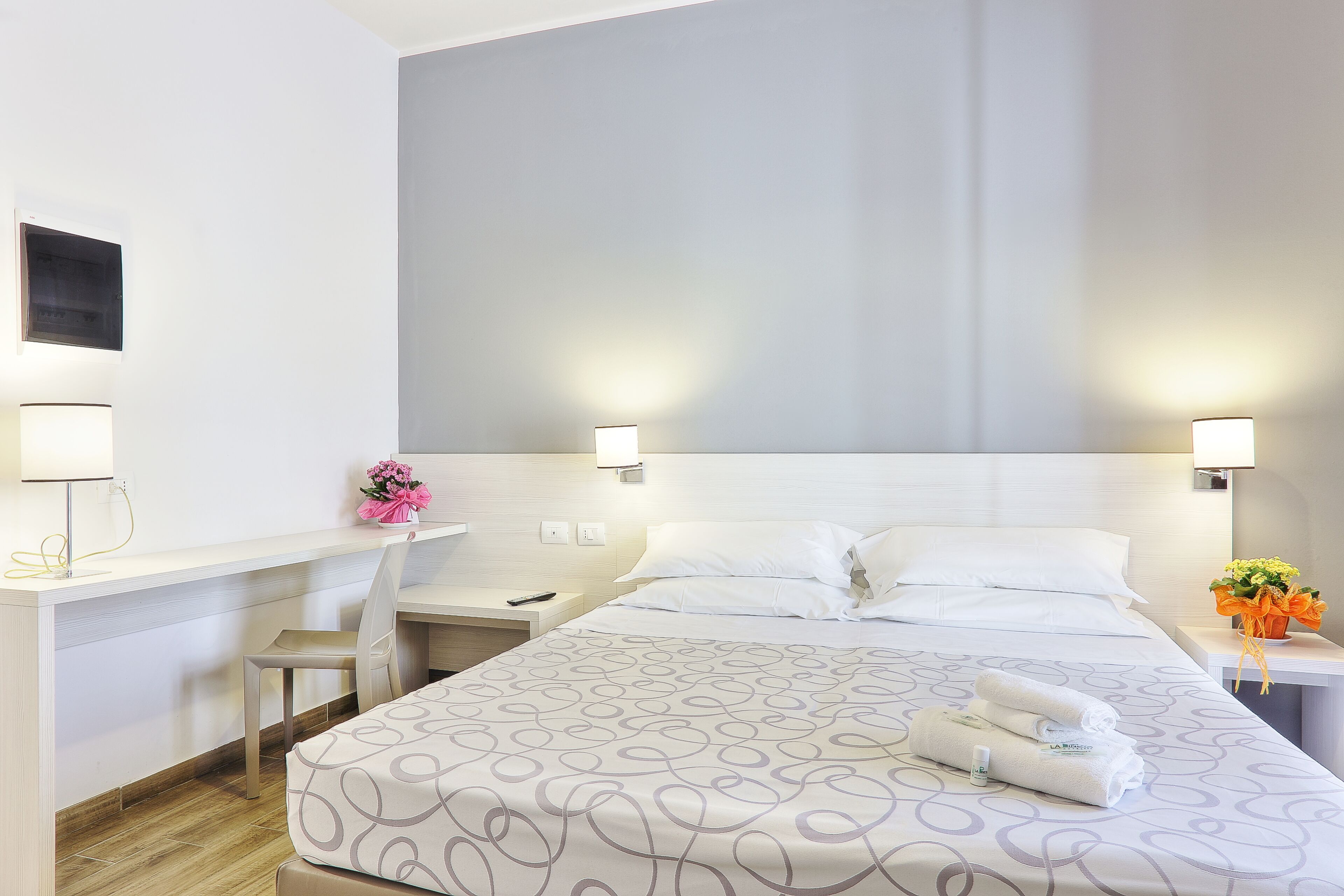 Habitació d'hotel amb un llit, un escriptori, una cadira i una tauleta de nit amb una làmpada.