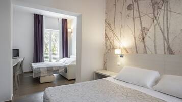 Habitación de hotel con cama, escritorio y silla. Se observa una ventana con cortinas, una lámpara de pared y un cuadro en la pared.