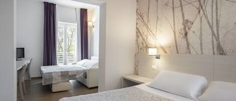 A hotel room with a bed, a desk, and a chair. There is a window with curtains, a wall-mounted light, and a painting on the wall.