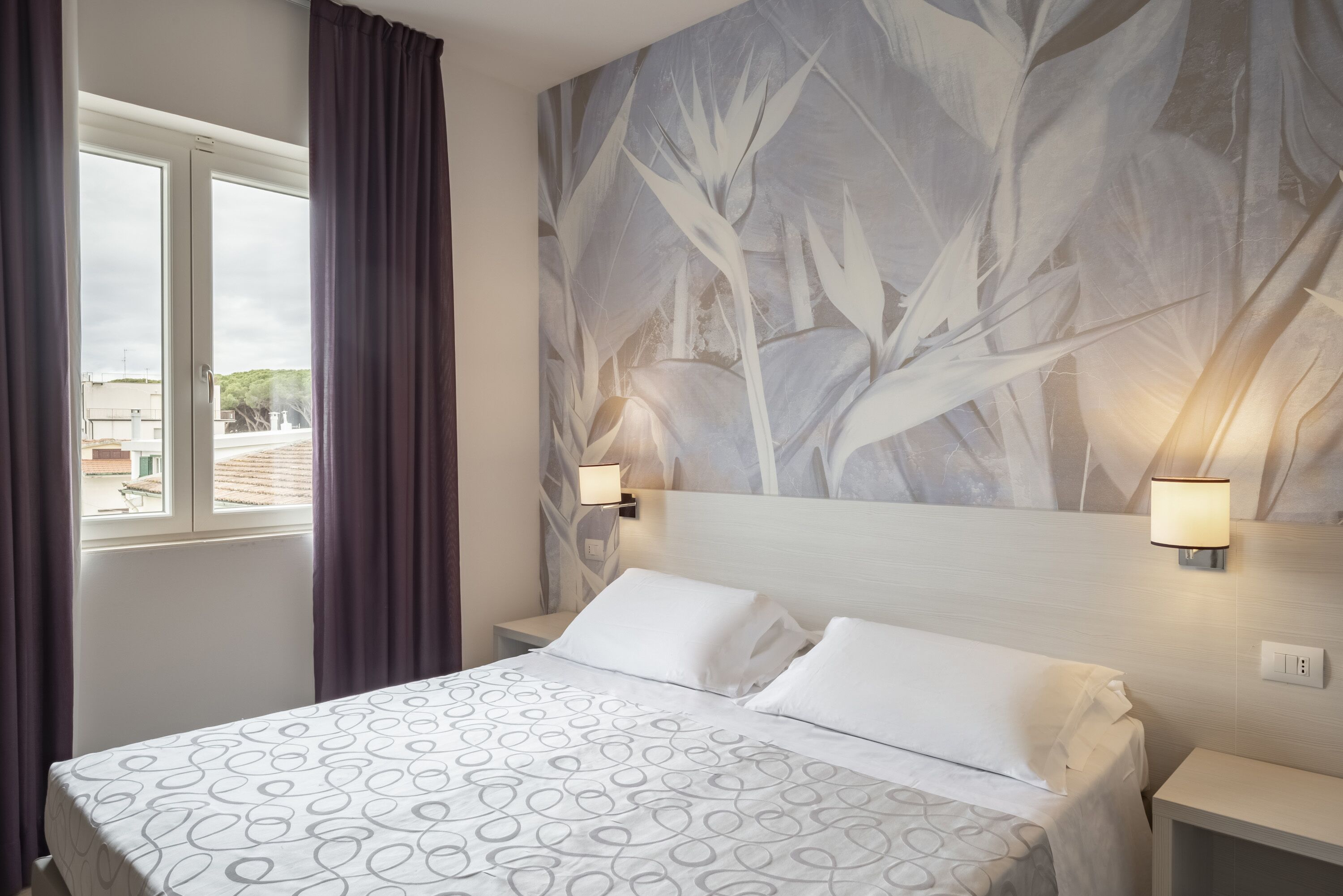 A hotel room with a bed, a large wall mural, two bedside lamps, and a window with curtains.