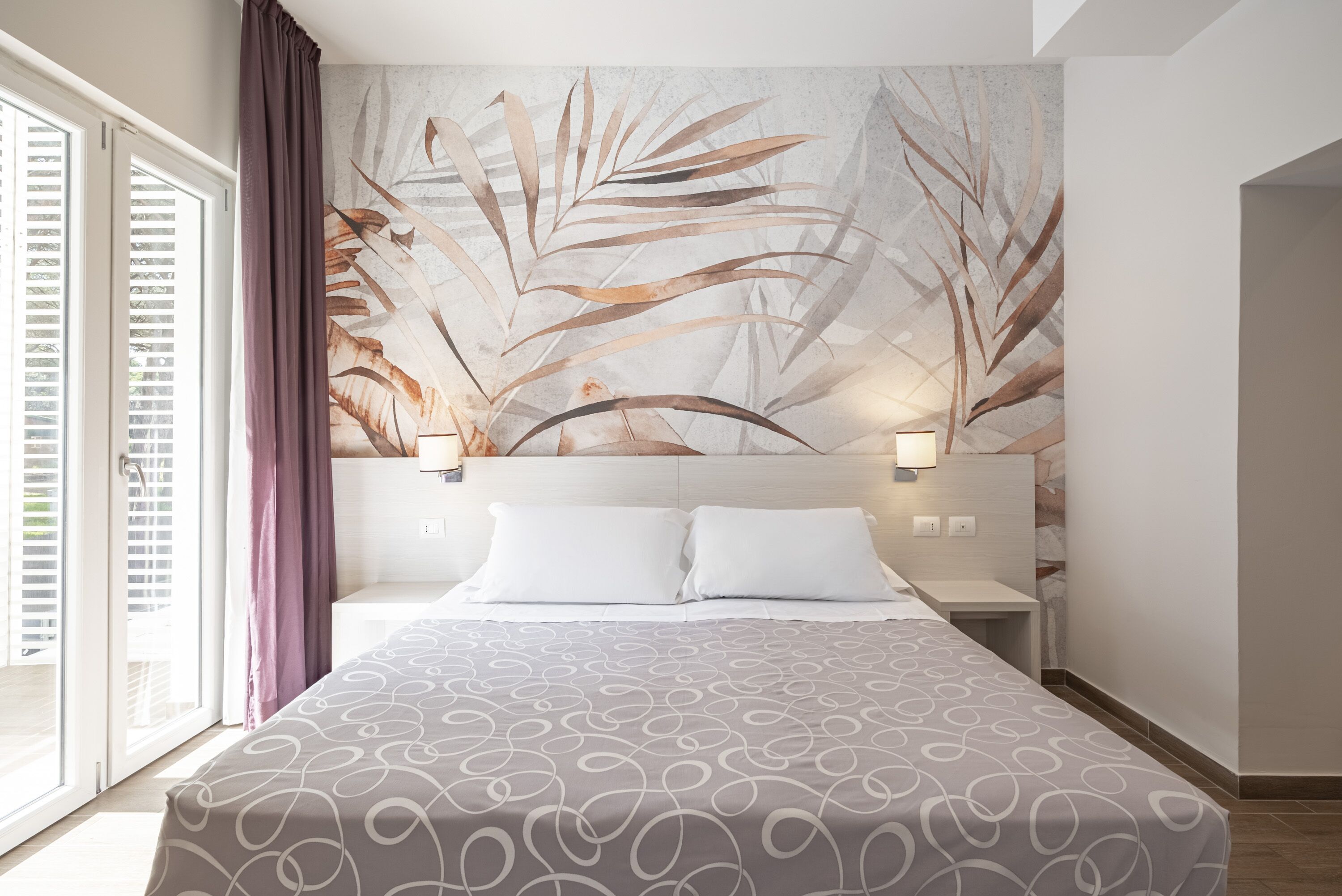 A hotel room with a bed, a large wall mural, and a window with curtains.