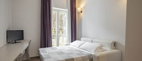 Ein Schlafzimmer mit einem Bett, einem Schreibtisch, einem Fernseher und einem Fenster mit Vorhängen.