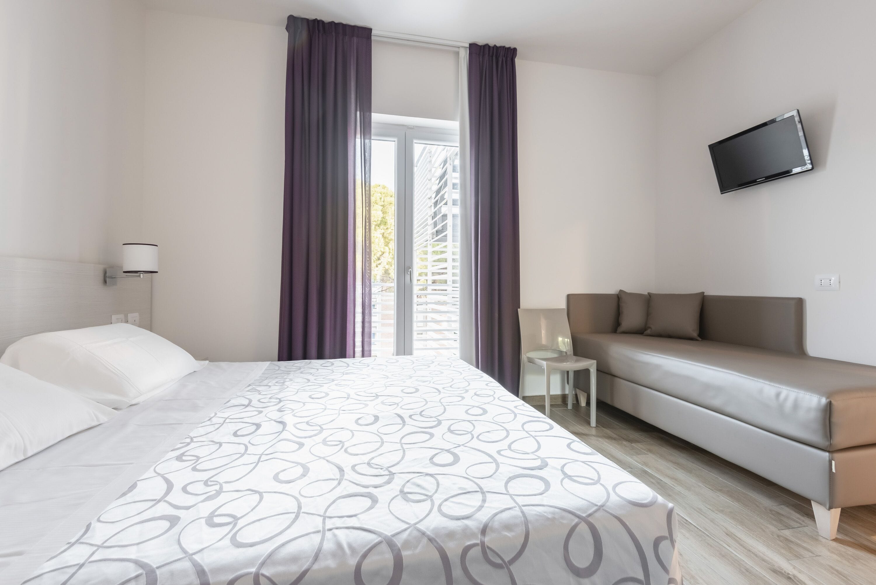 A hotel room with a bed, a sofa, a TV, and a window with curtains.