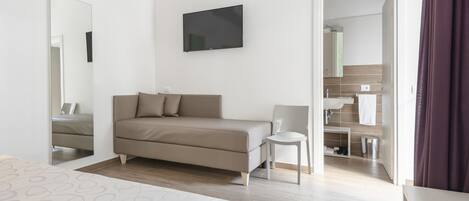 A modern hotel room with a bed, a sofa, a TV, and a bathroom visible through an open door.