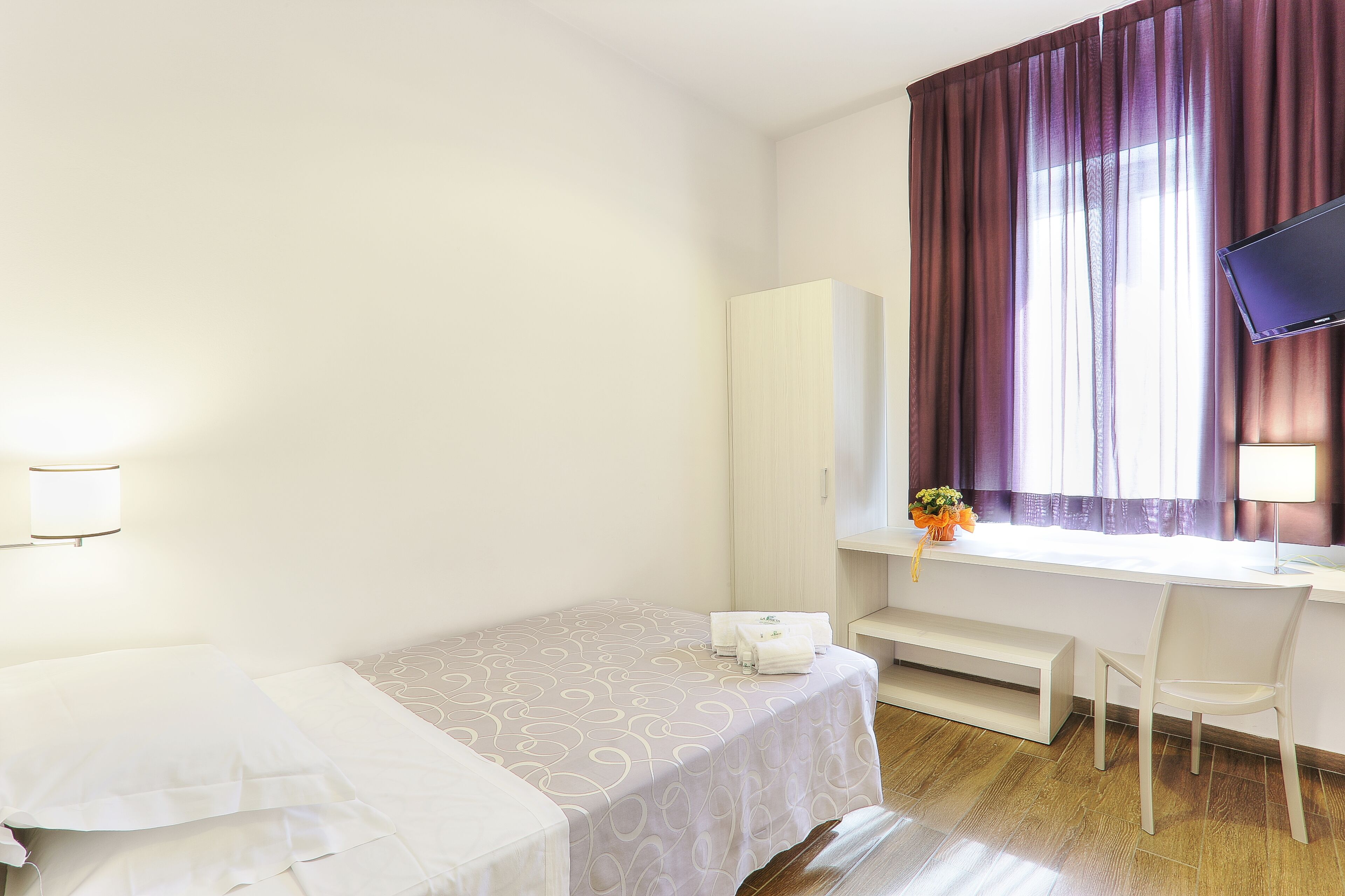 A hotel room with a bed, a desk with a chair, a television, and a wardrobe.