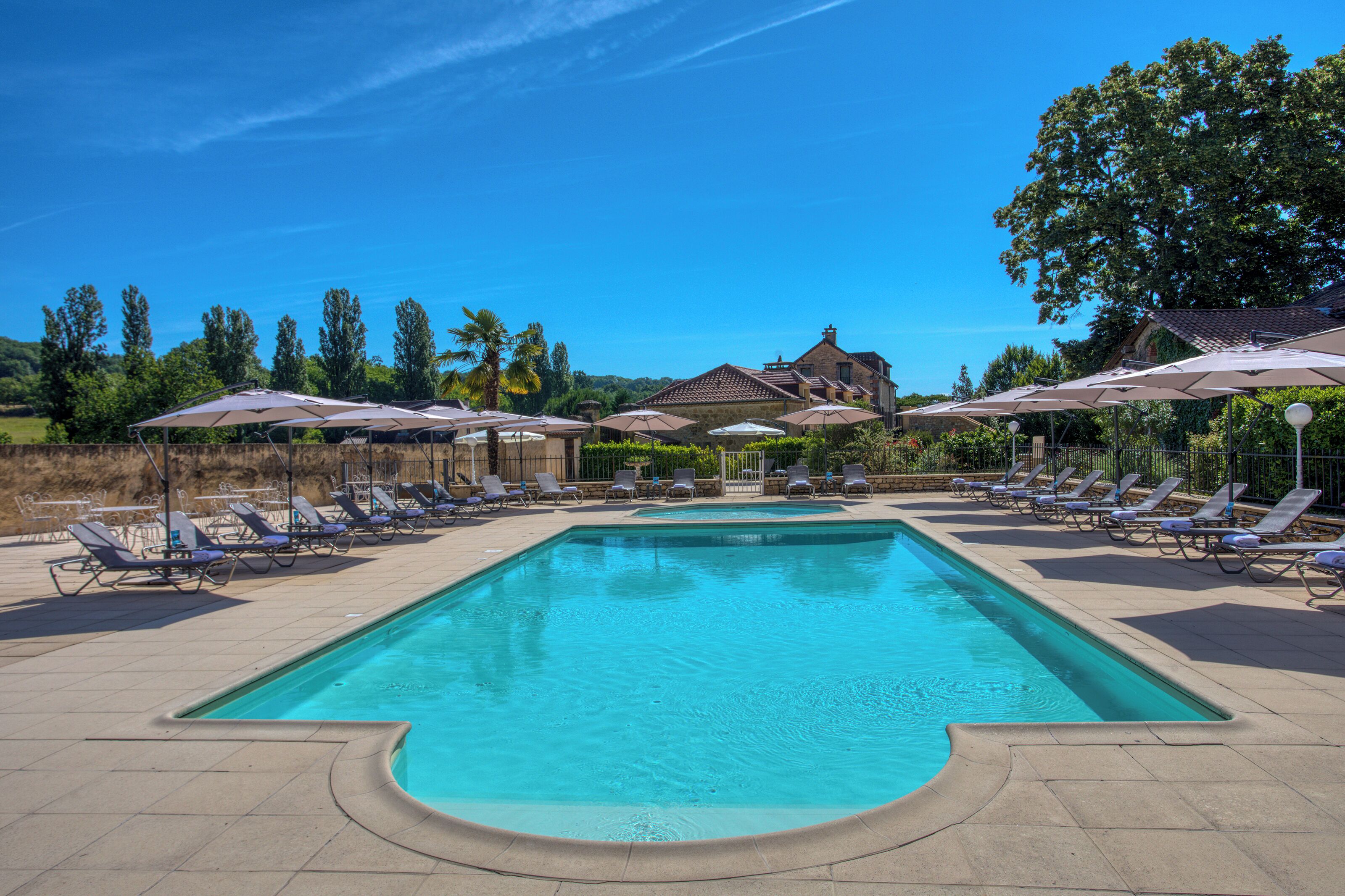 Une piscine entourée d’arbres, avec des chaises longues et des parasols, et un bâtiment visible au loin.