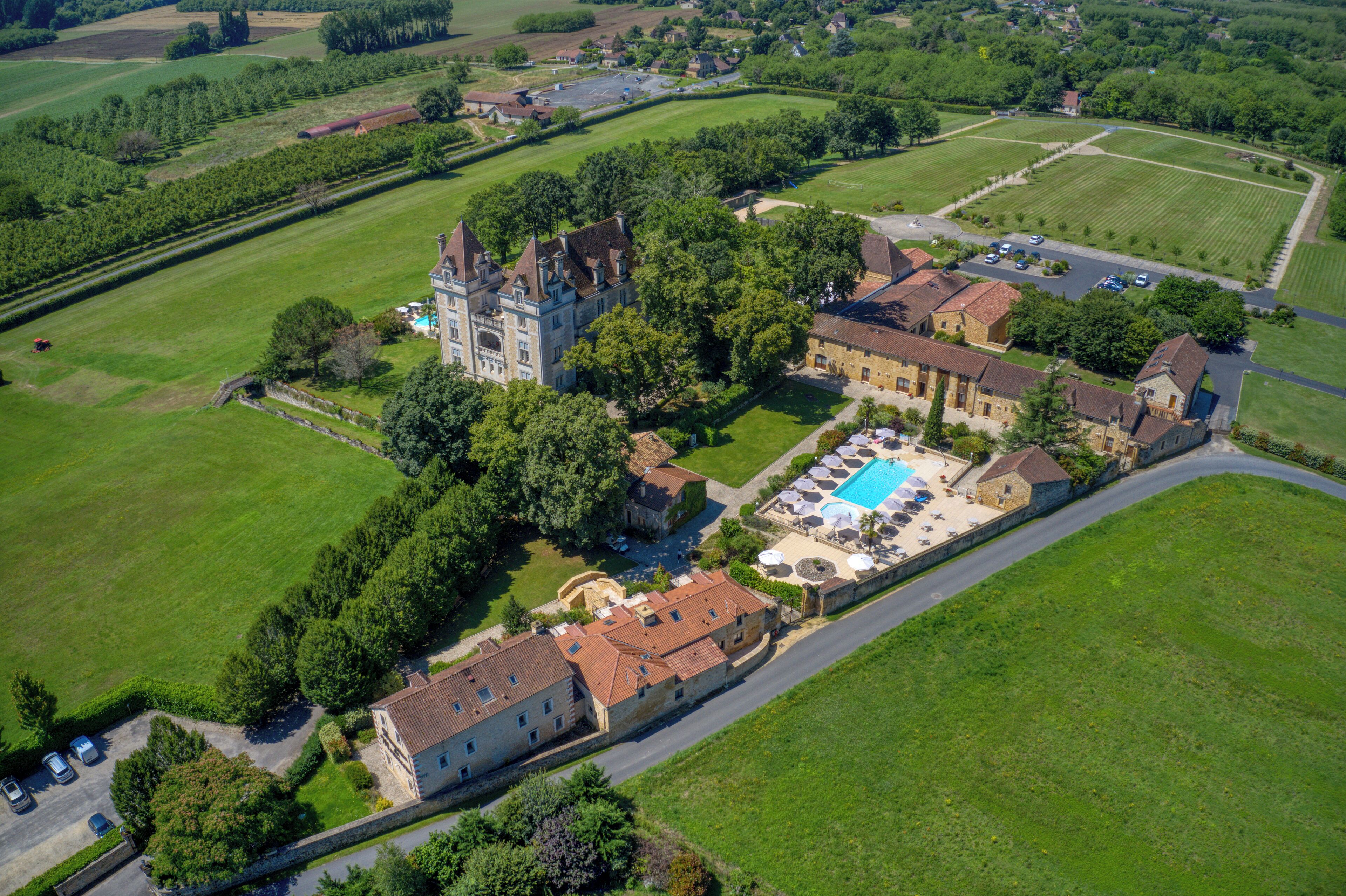 An aerial view of a large estate with a castle-like building, a swimming pool, and surrounding green fields.