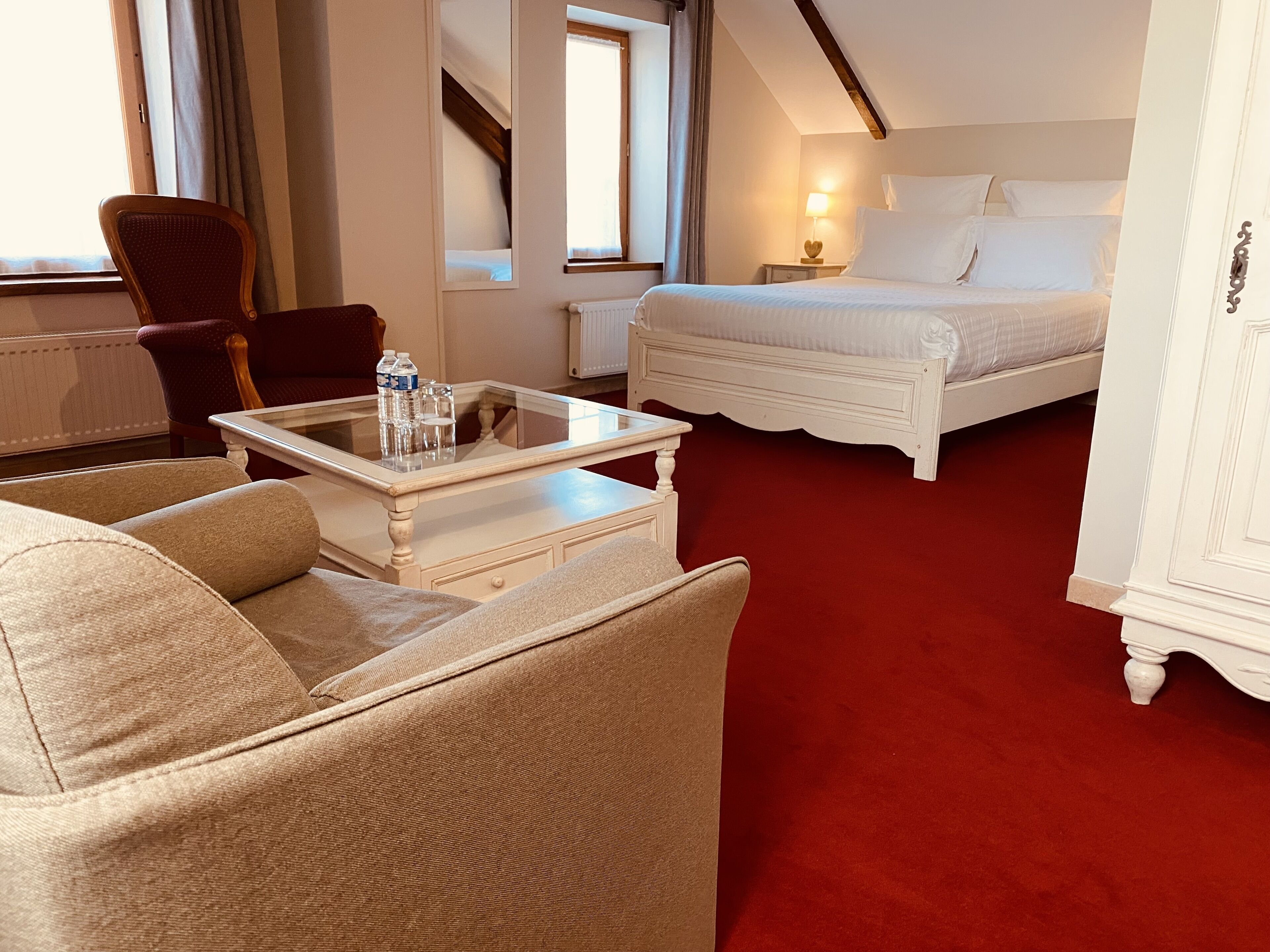 Une chambre d’hôtel avec un lit, une chaise, une table avec des bouteilles d’eau, une lampe et une fenêtre.