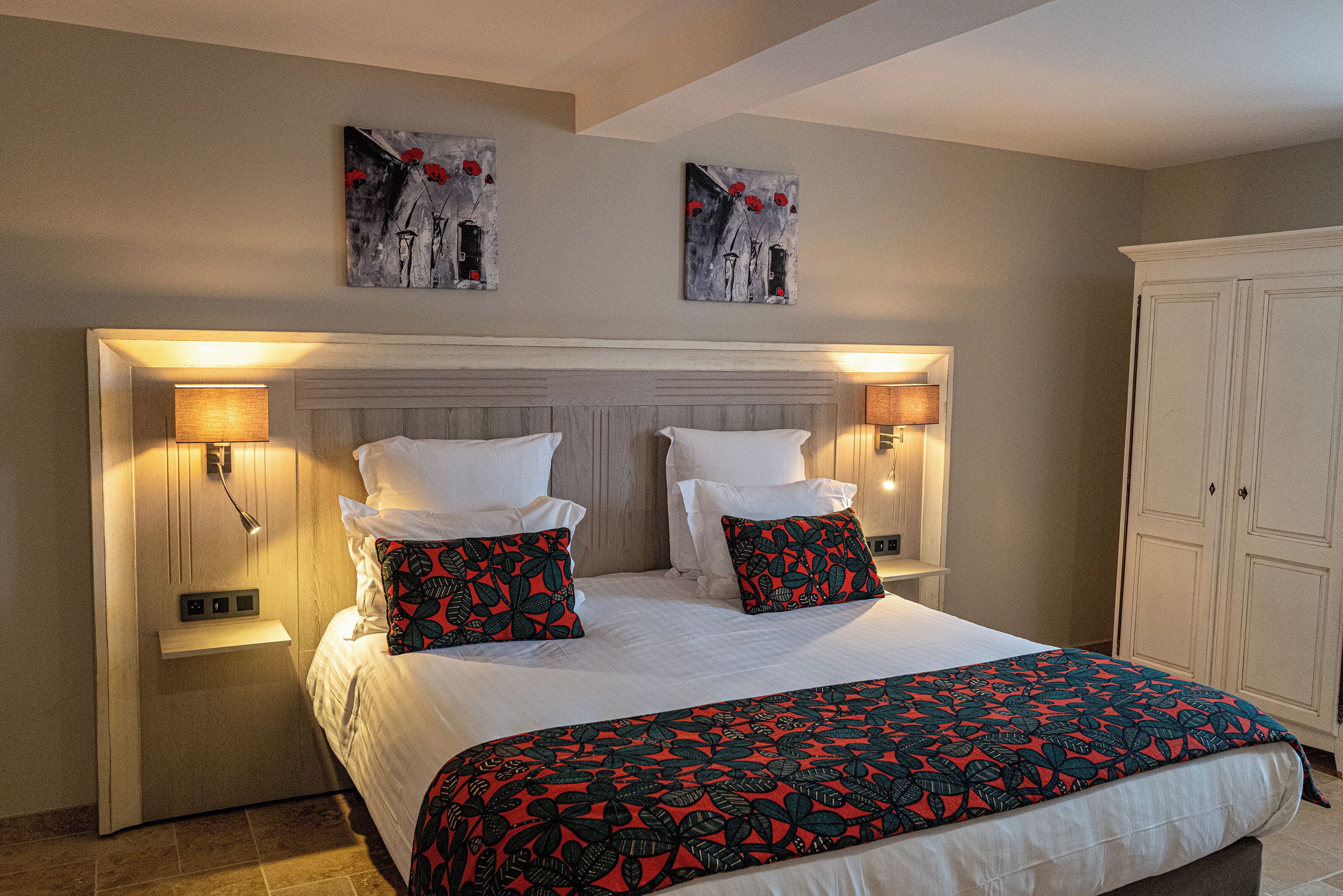 A double bed with a floral-patterned bedspread, two wall-mounted lamps, and two paintings above the bed.