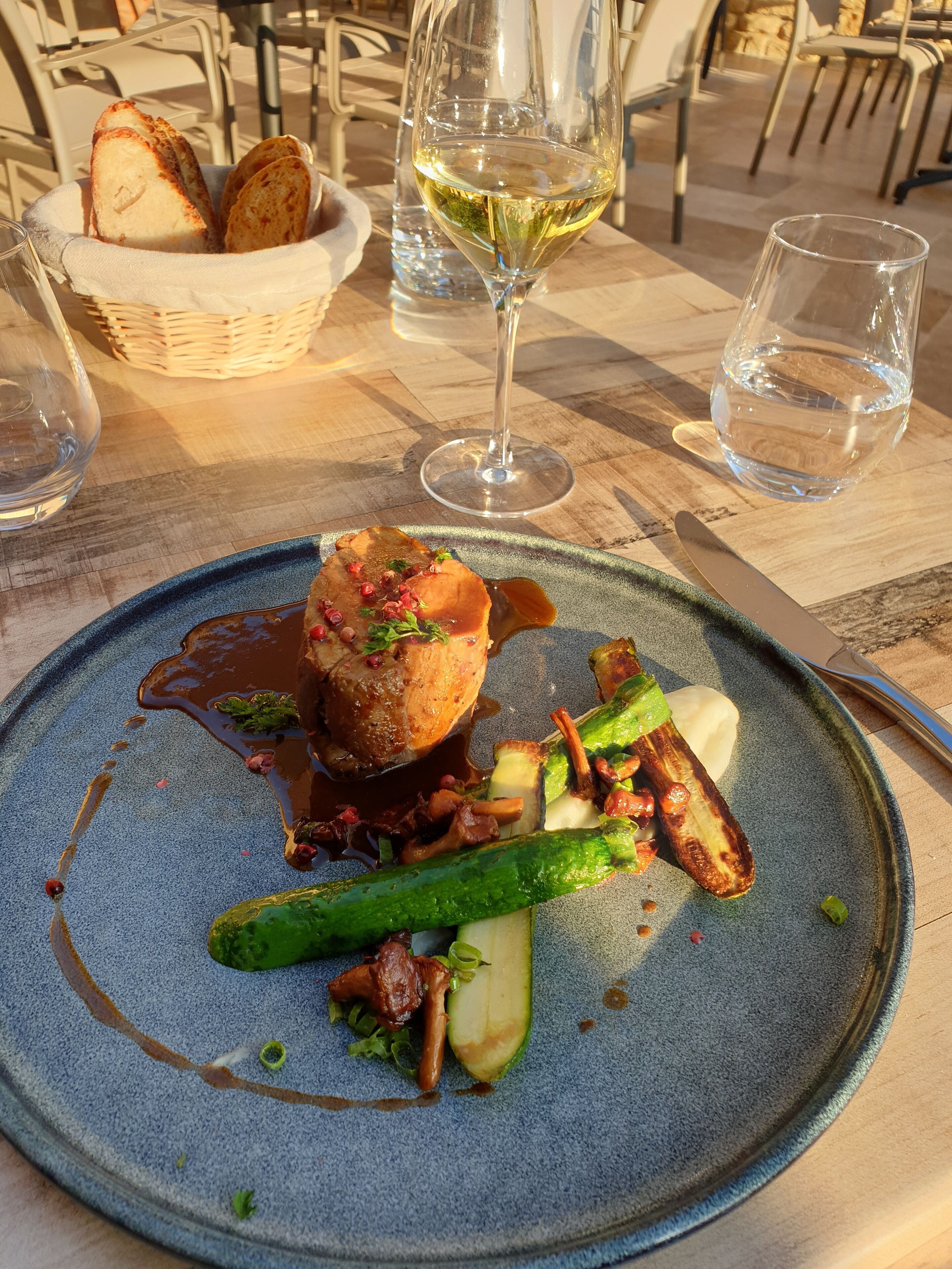 A dining table with a plate of food, a glass of white wine, and a basket of bread.