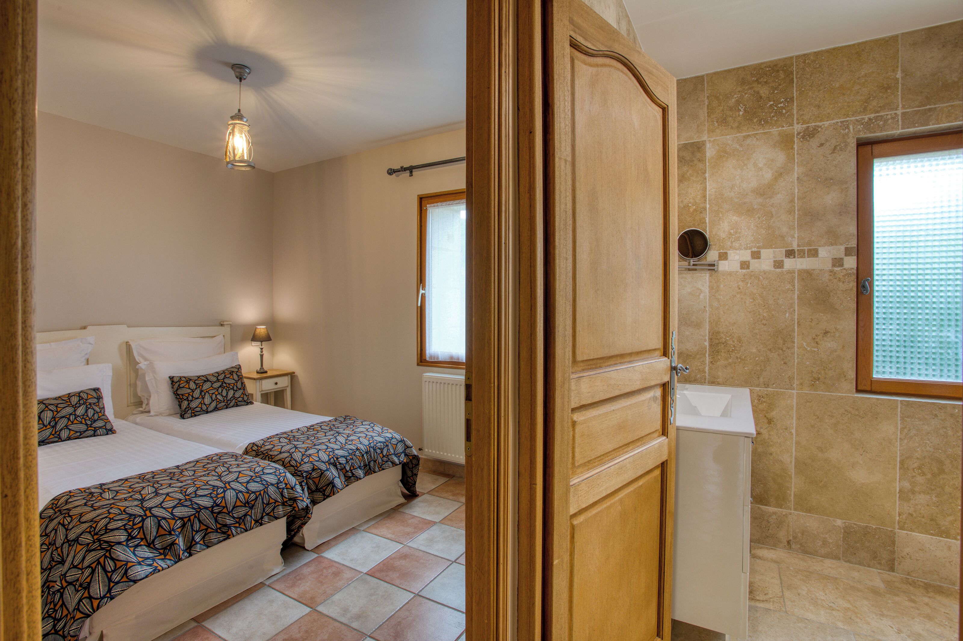 A bedroom with two beds, a nightstand, a lamp, and a bathroom with a shower and a window.