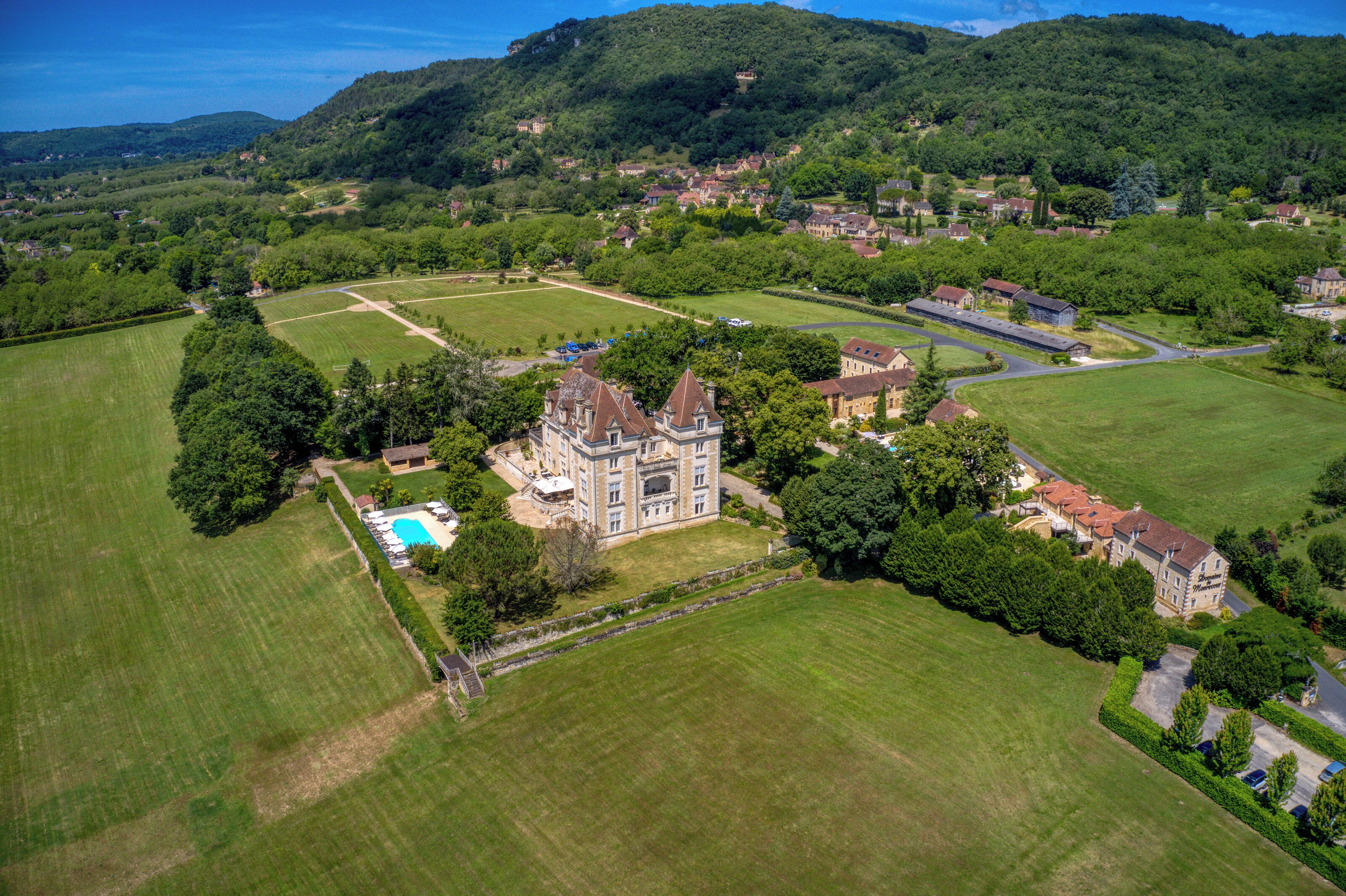 Vista aérea de una extensa propiedad con un imponente edificio parecido a un castillo, amplios campos verdes y una piscina.
