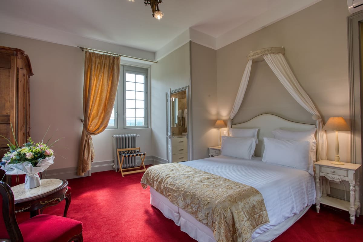 A bedroom with a four-poster bed, a bedside table with a lamp, a chair, a dresser, and a window with curtains.