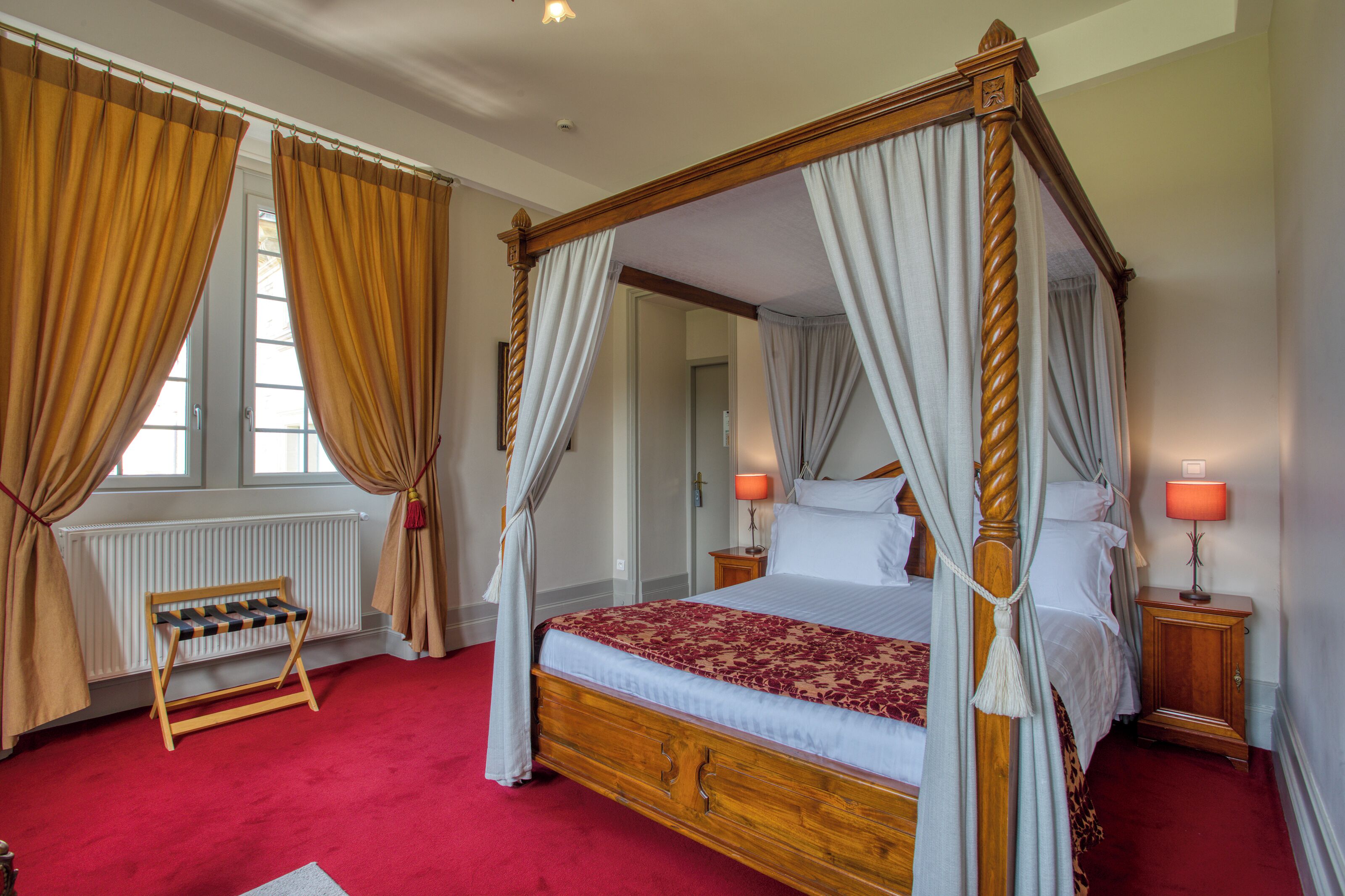 A four-poster canopy bed with a red and white bedspread, a wooden nightstand with a lamp, and a small bench with a chessboard.