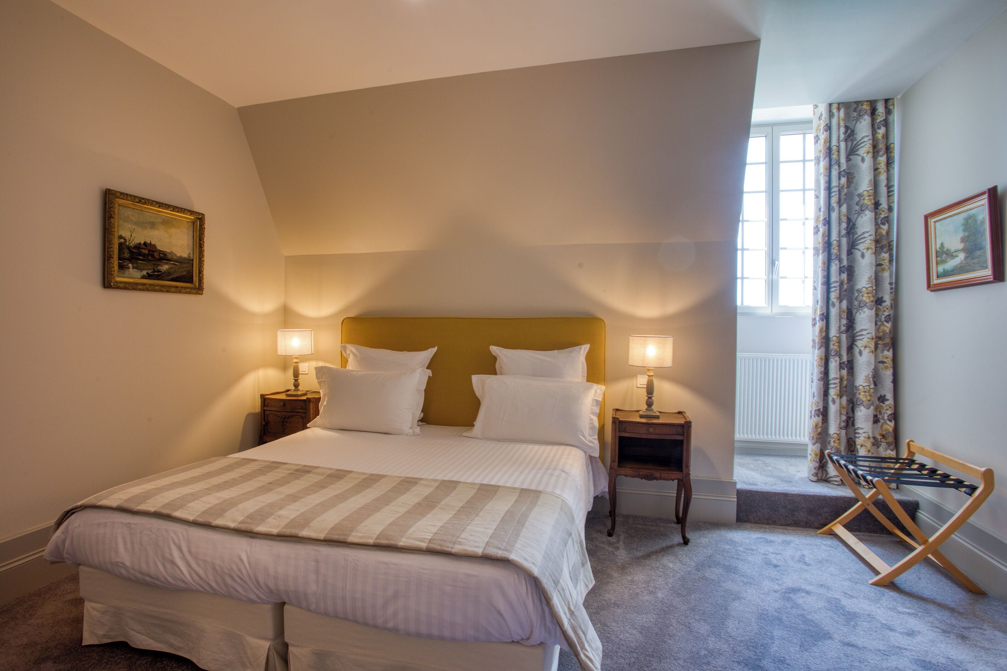 Une chambre à coucher avec un lit, deux tables de chevet avec des lampes, une fenêtre avec des rideaux et un tableau au mur.