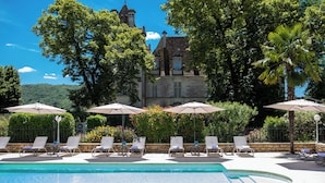 A pool area with lounge chairs and umbrellas, surrounded by lush greenery and a historic building in the background. - Domaine du Château de Monrecour – Hôtel et Restaurant (Saint-Vincent-de-Cosse)