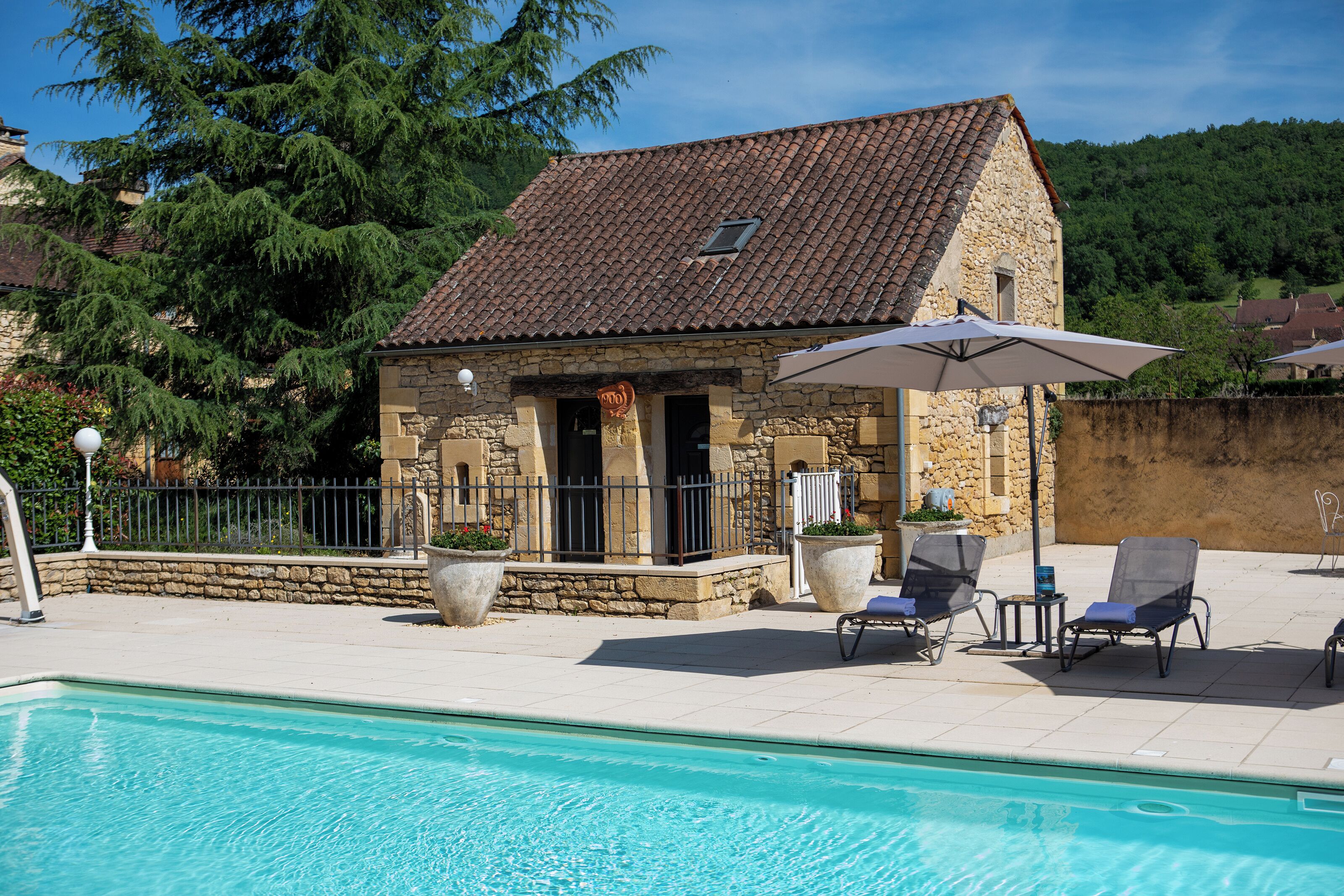 A stone building with a tiled roof, a swimming pool, and outdoor seating with umbrellas.