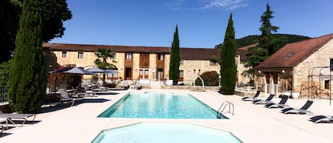 A pool area with lounge chairs and umbrellas, surrounded by stone buildings with tiled roofs.